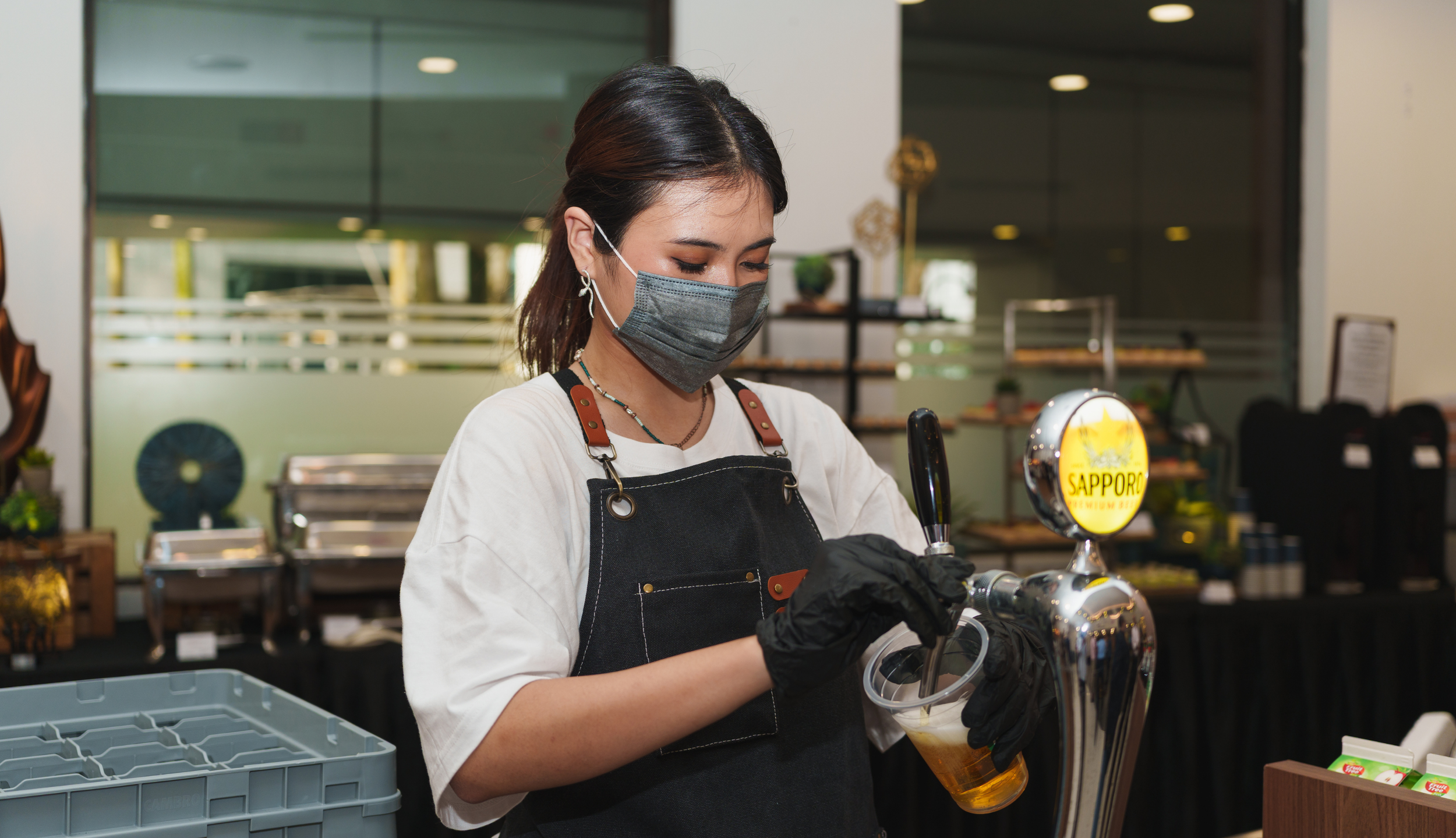 A woman wearing a face mask, black gloves, and an apron pouring beer from a tap into a plastic cup in a modern indoor setting.