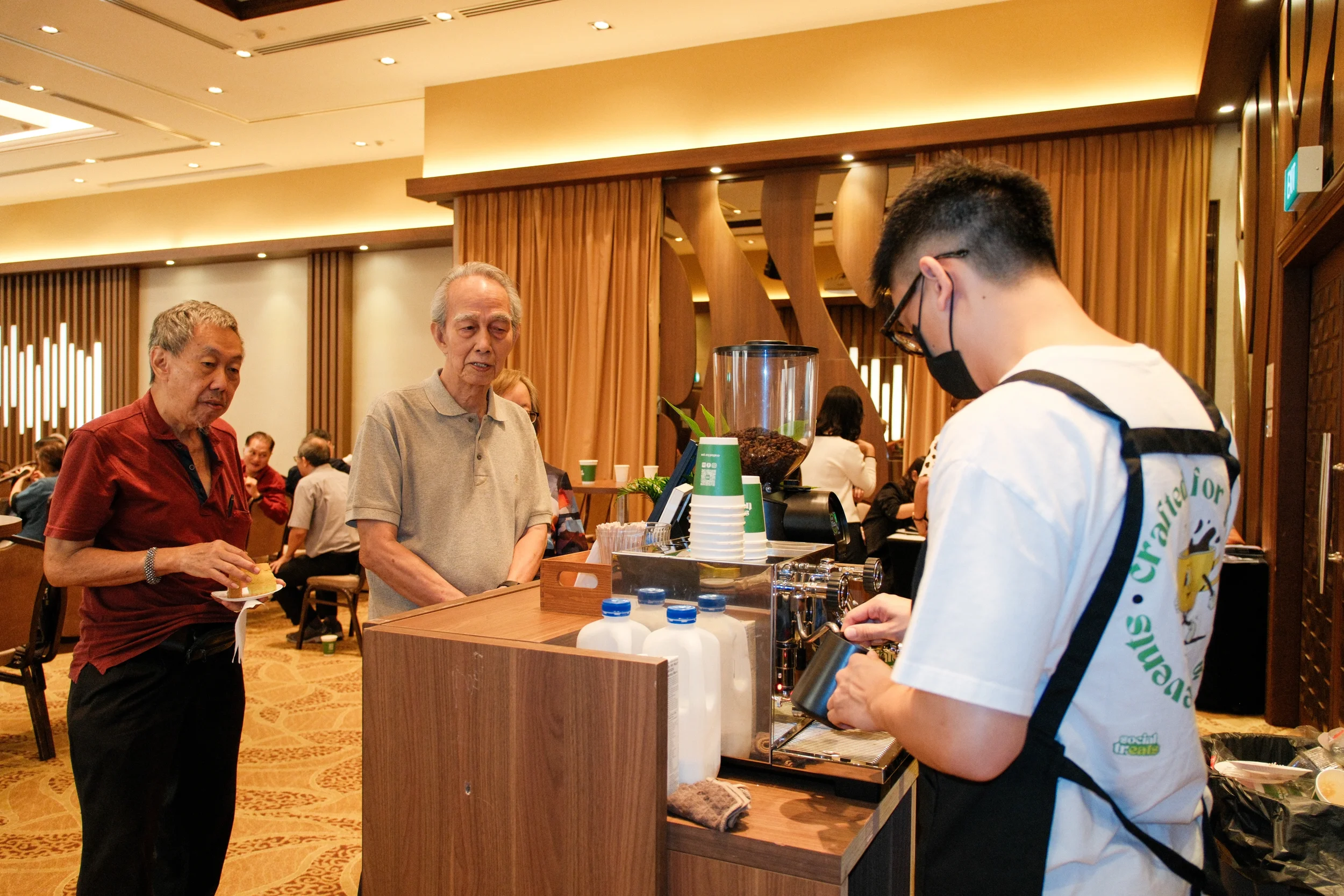 A barista wearing a white shirt, glasses and a black face mask making coffee at a coffee station in a busy indoor cafe or restaurant with customers in the background.