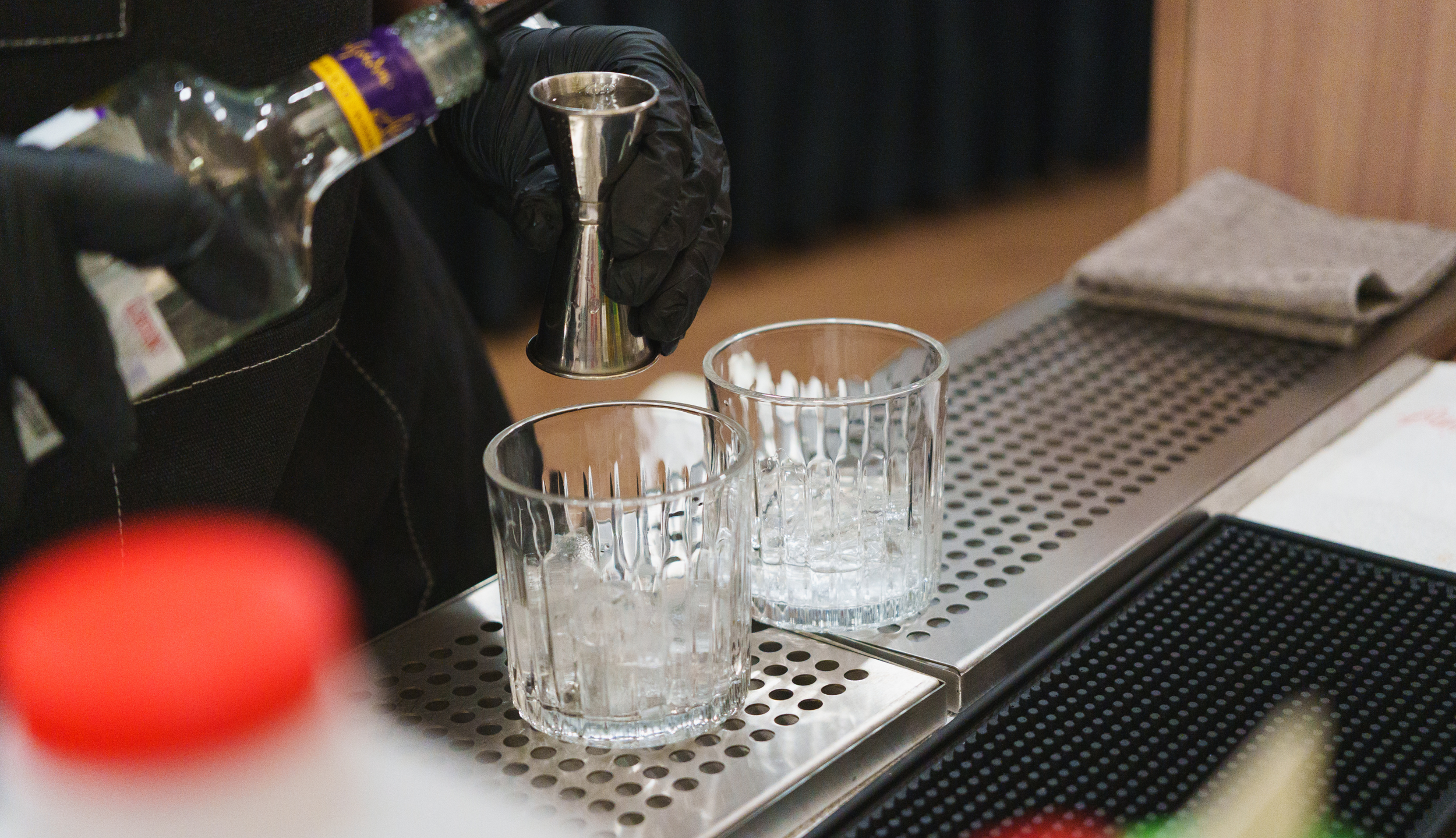 Person wearing black gloves pouring liquid from a bottle into a jigger, two glasses with ice on a bar counter, bar tools and towel in the background.