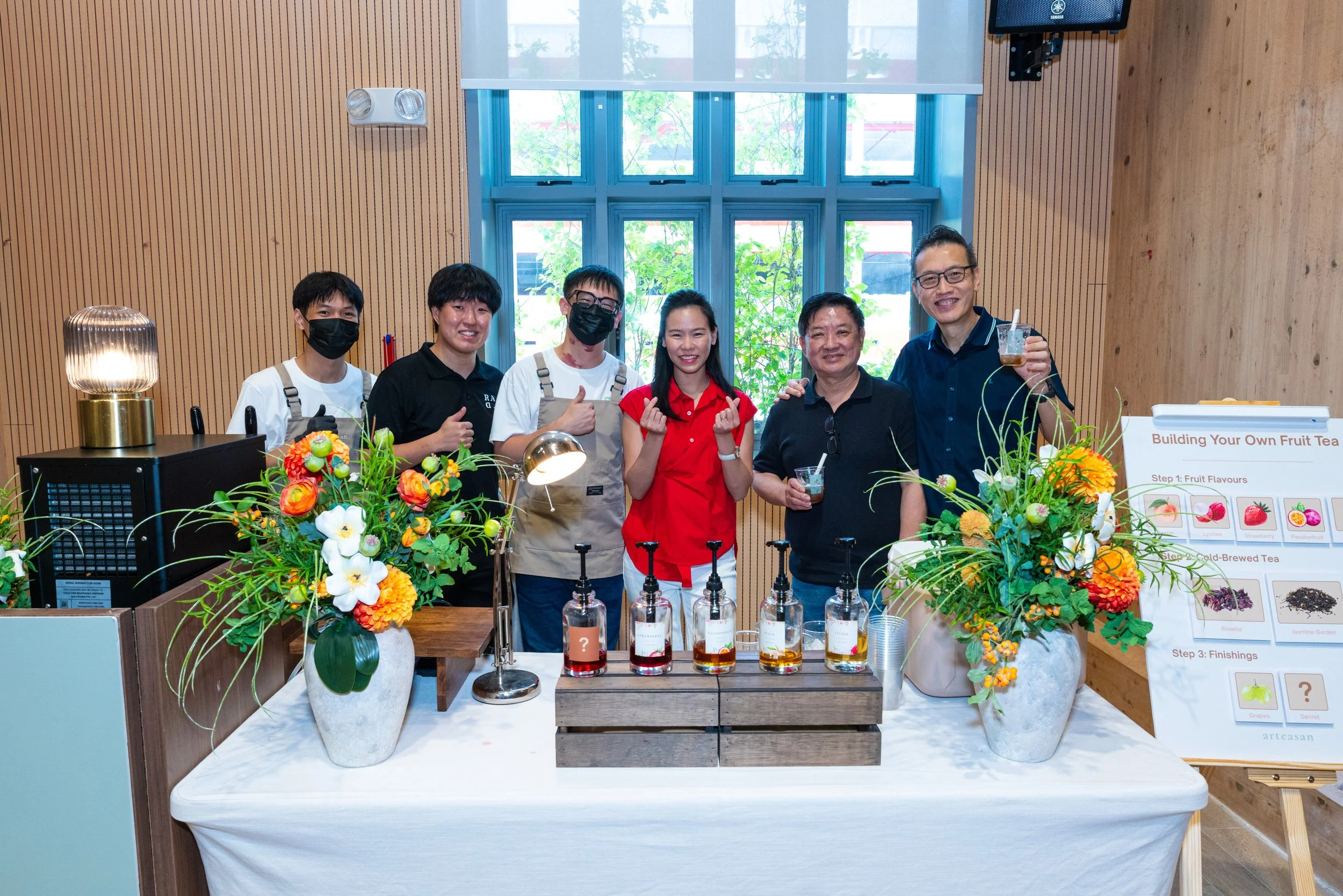 Group of six people standing behind a table with colorful flower arrangements and bottles of ingredients, smiling and holding drinks, in a bright room with large windows, wooden walls, and a sign about building fruit tea.