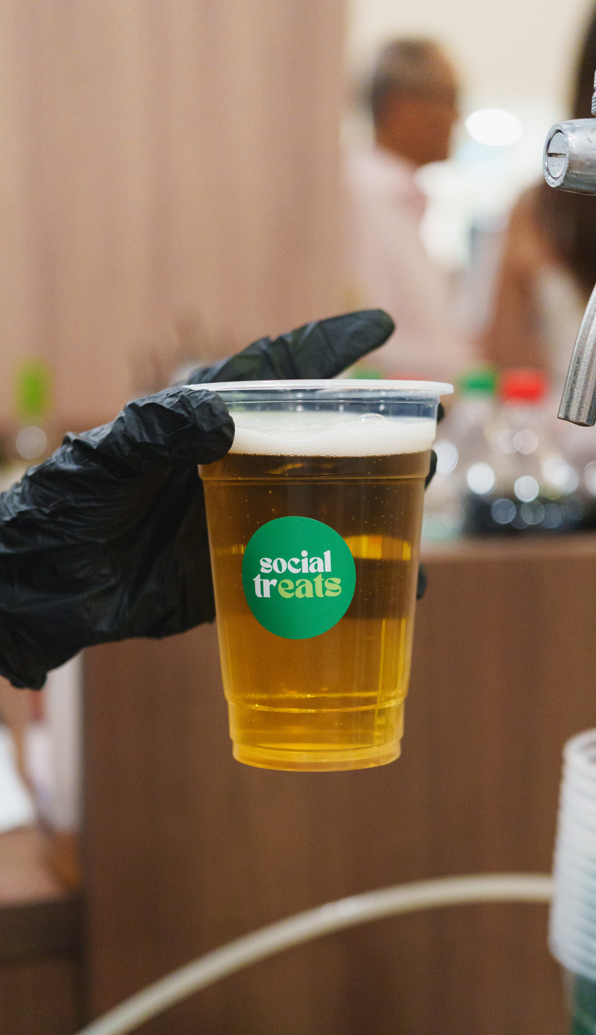 A hand in a black glove holding a clear plastic cup of beer with a green circular sticker that reads 'social treats'; the background is blurred with people and objects.