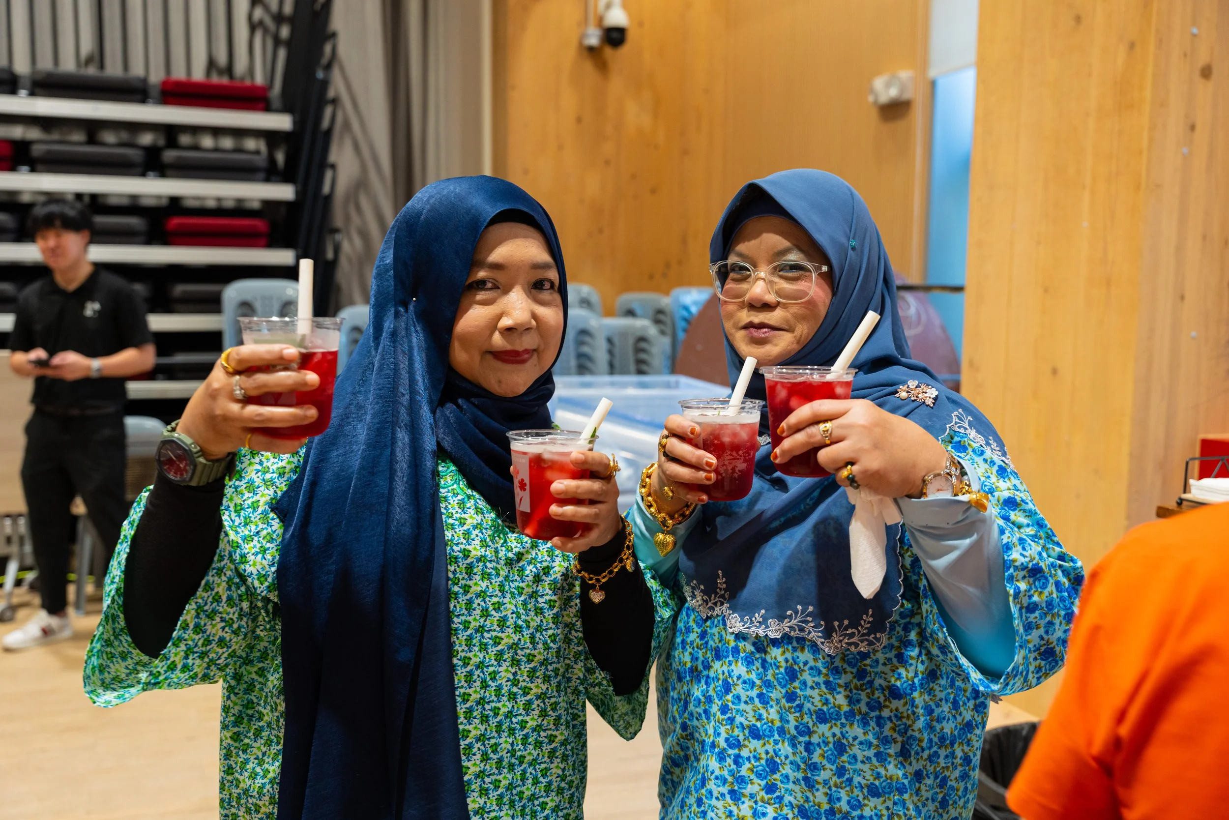 Two women wearing floral blouses and blue hijabs holding drinks with straws, celebrating indoors.