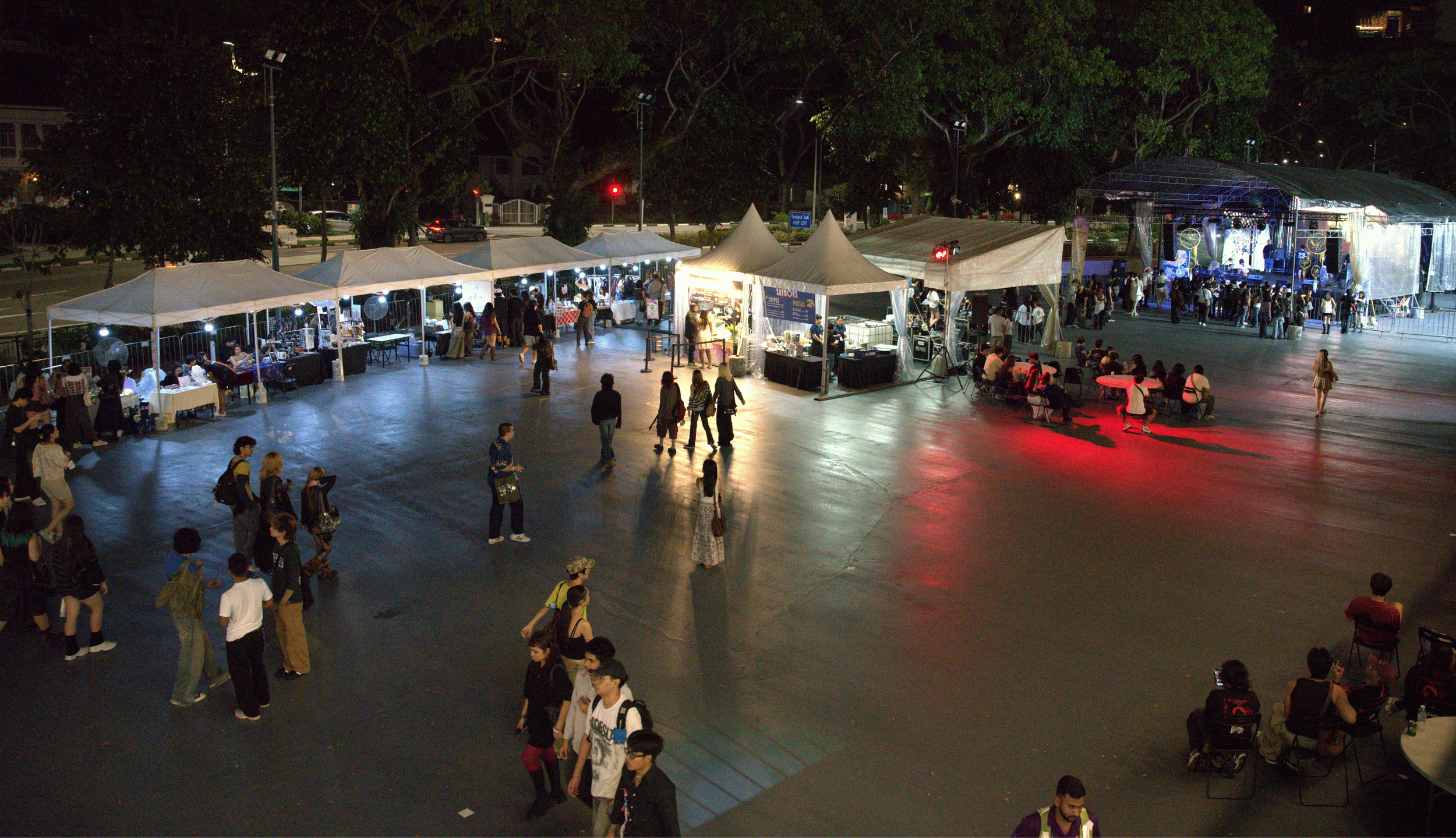 Nighttime outdoor event with food stalls and a stage. People walking and sitting around, trees in the background.