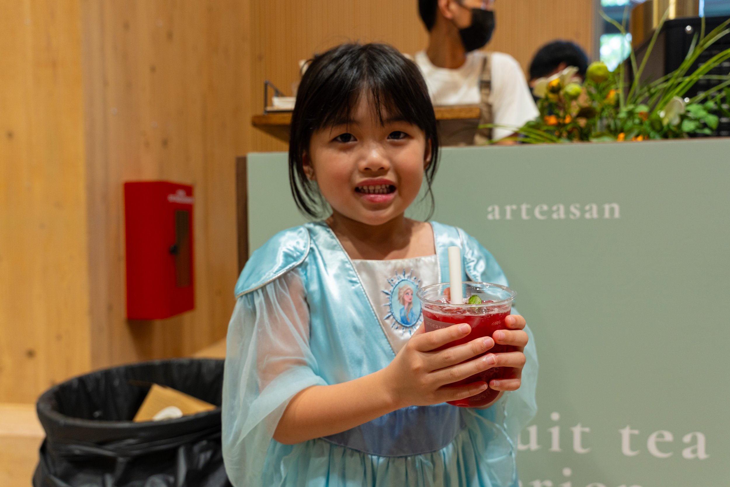 Young girl dressed as Elsa from Frozen holding a red fruit drink with a straw in a store or cafe.
