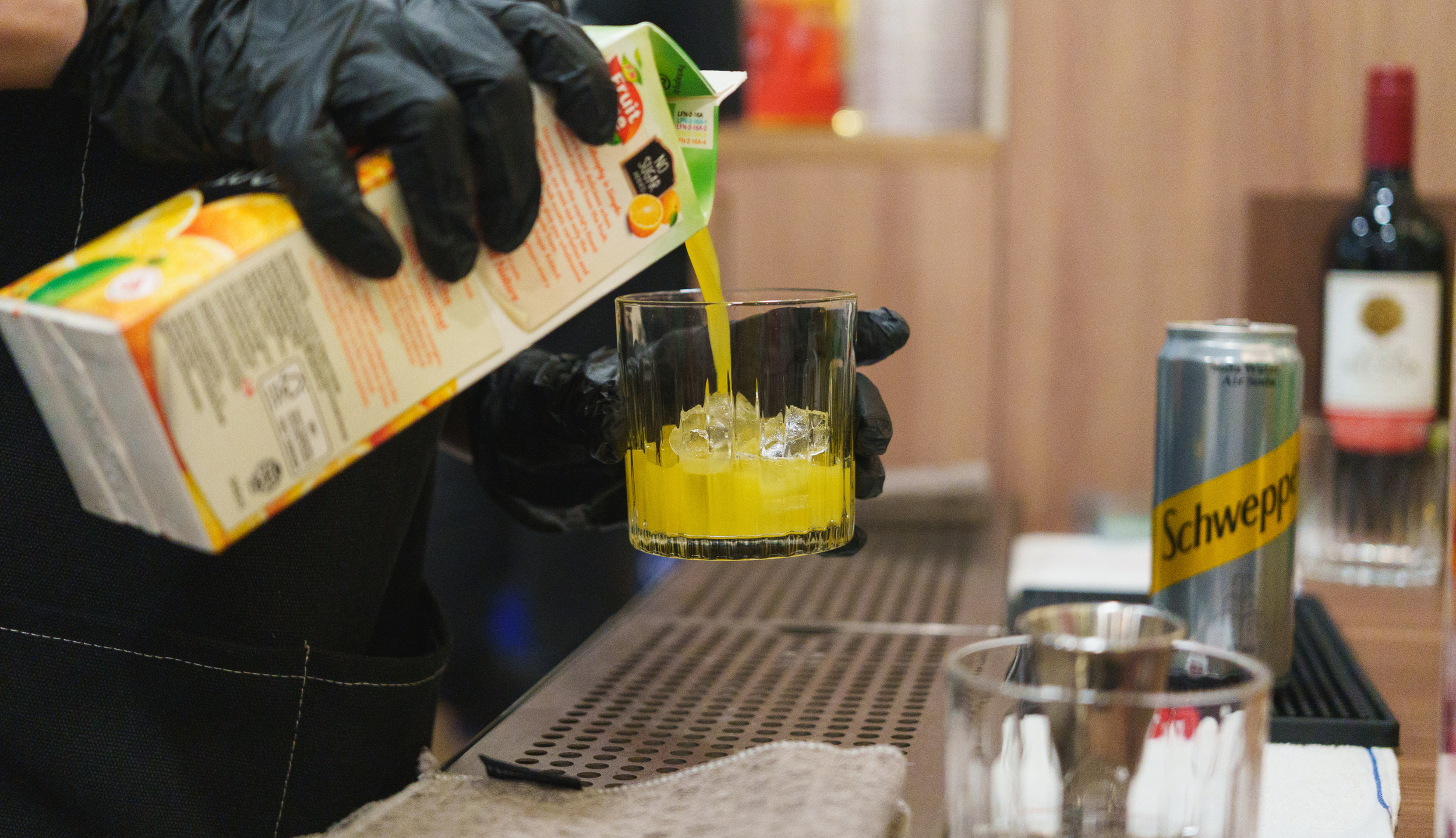 Person pouring orange juice into a glass with ice, wearing black gloves, with a can of Schweppes and a bottle of wine in the background.
