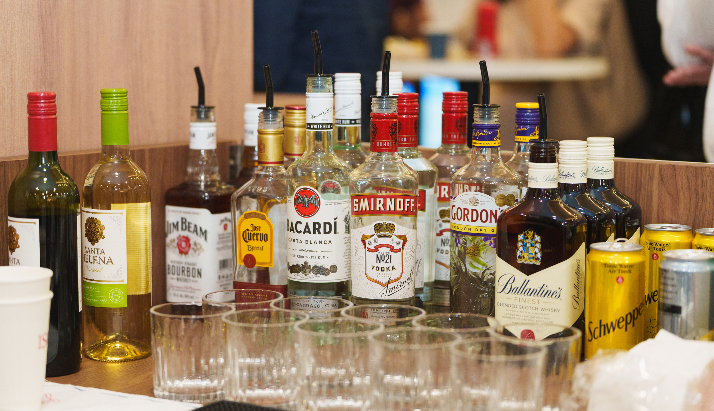 Various bottles of alcoholic beverages, including vodka, gin, and whiskey, are arranged on a bar counter with some empty glasses and canned drinks in the foreground.