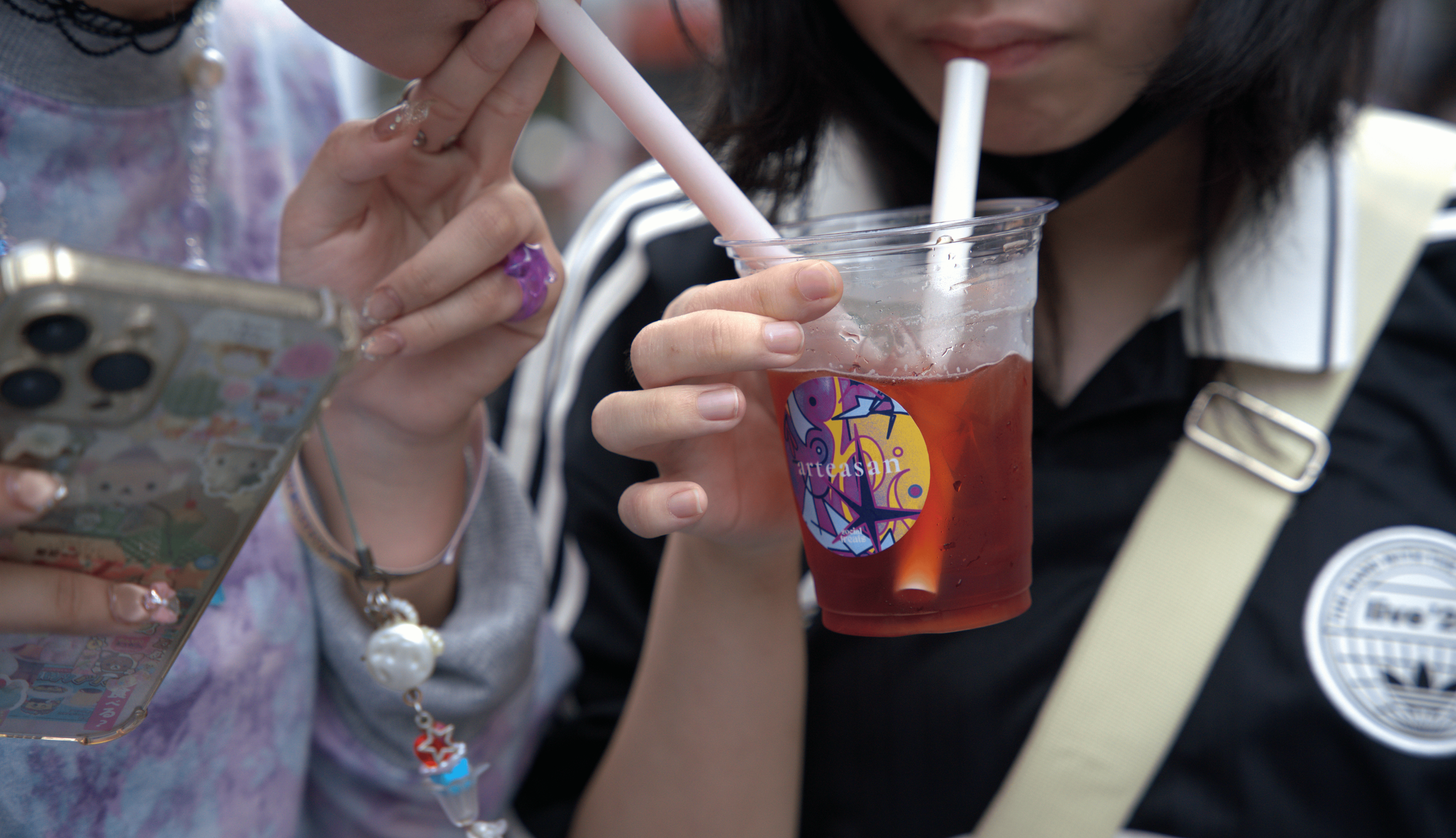 Two women, one drinking from a clear plastic cup with a straw, the other holding a smartphone decorated with stickers.