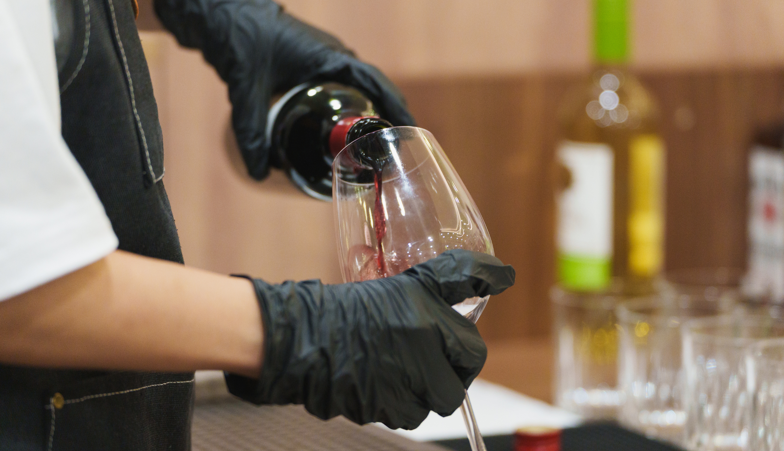 Person pouring red wine from a bottle into a glass, wearing black gloves.