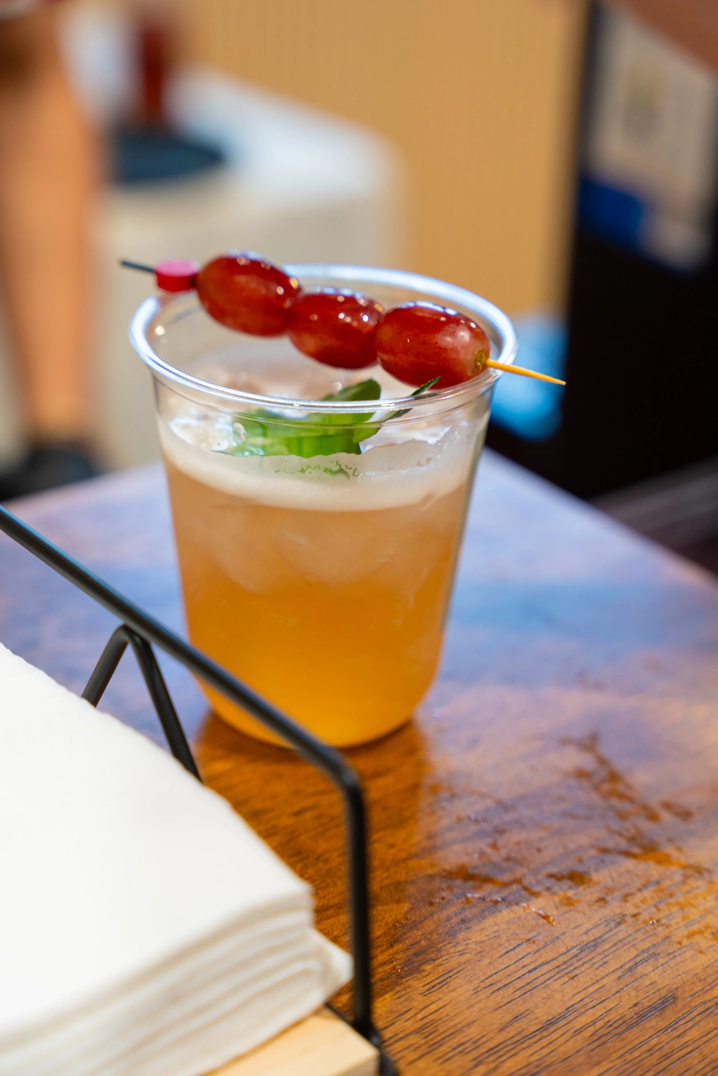 A cocktail glass with a yellow-orange drink, garnished with green mint leaves and a skewer of red grapes on top, on a wooden table with a napkin holder nearby.