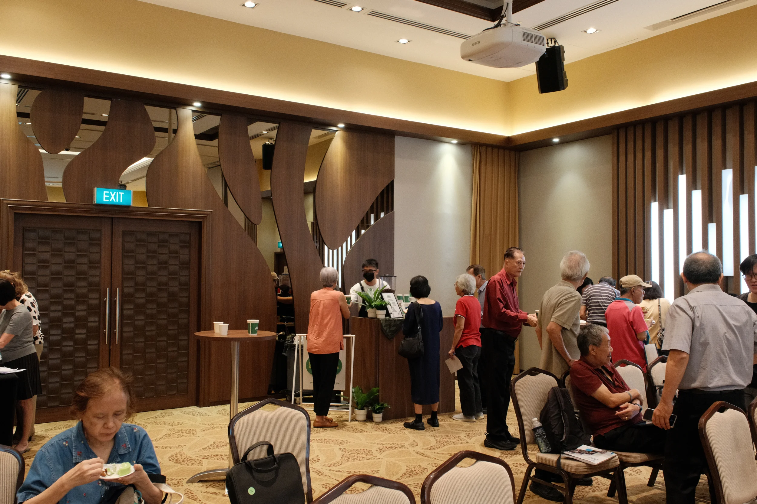Indoor scene in a cafe or waiting area with elderly and middle-aged people standing in line and seated, some eating, during daytime. The room features wood paneling, curtains, and a ceiling-mounted projector, with an exit sign above a door.