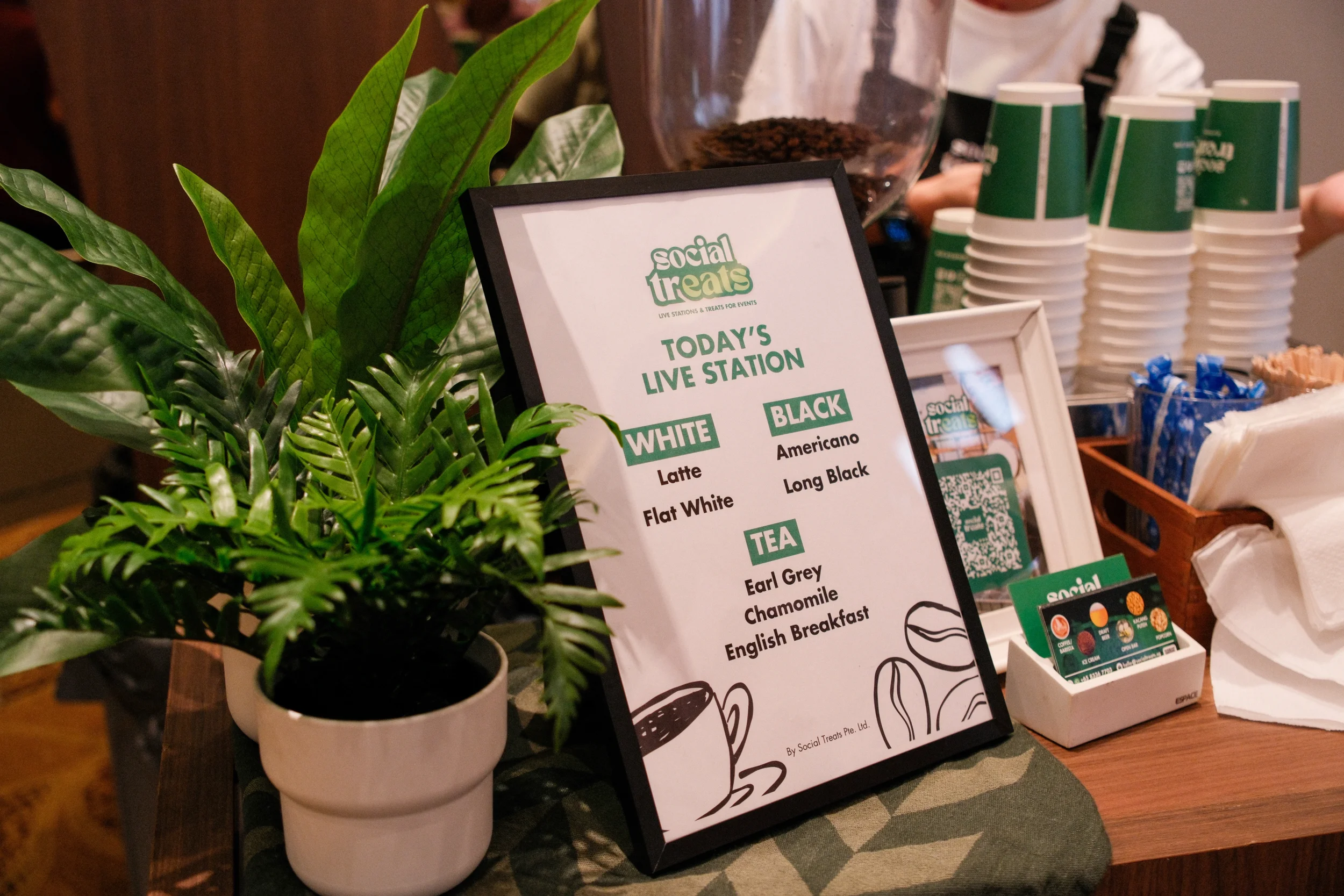 A table with a menu sign for Social Treats' live station, displaying drink options: white, black, and tea. A plant in a white pot, paper cups, sachets, and small framed photos are also on the table.