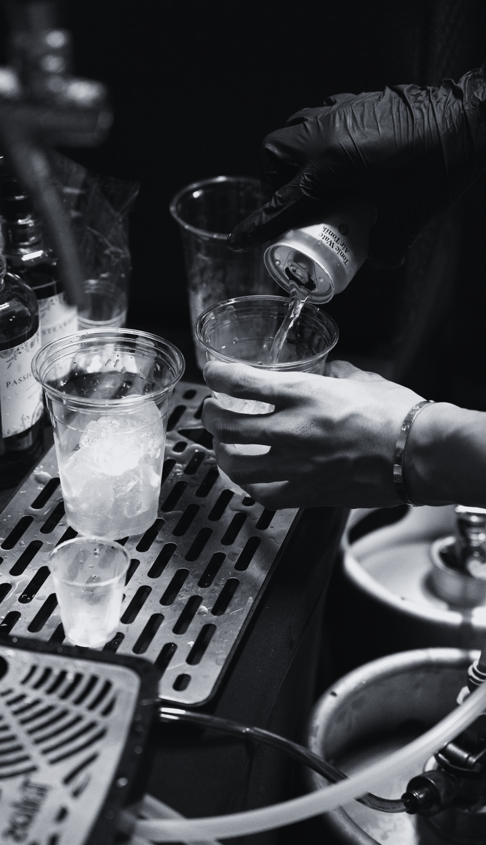 A person wearing black gloves pours a clear beverage into a plastic cup with ice, on a bar counter with other cups and bottles around.
