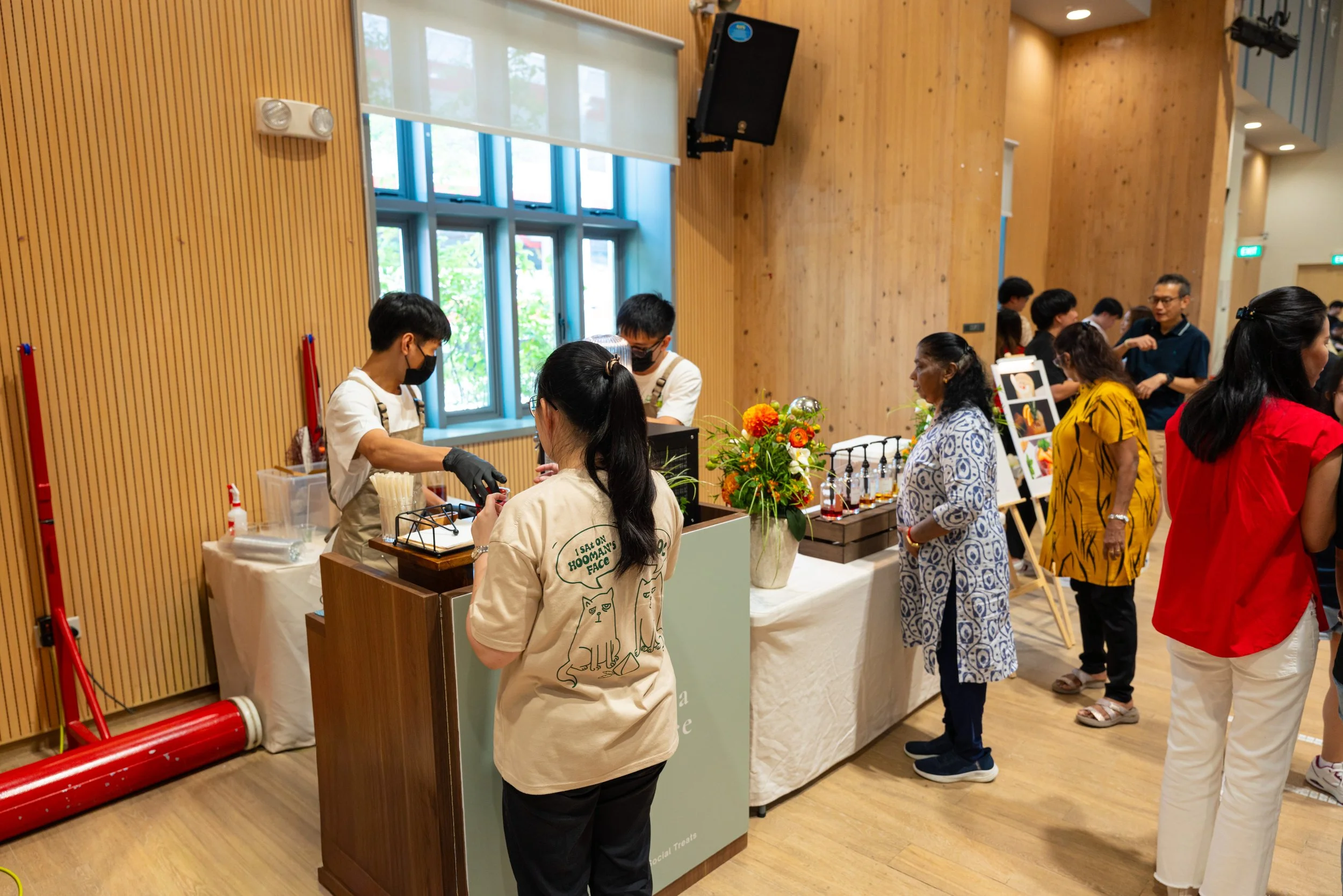 People at a market or event standing in line at a booth with flowers, bottles, and other items, inside a room with wooden walls and windows.