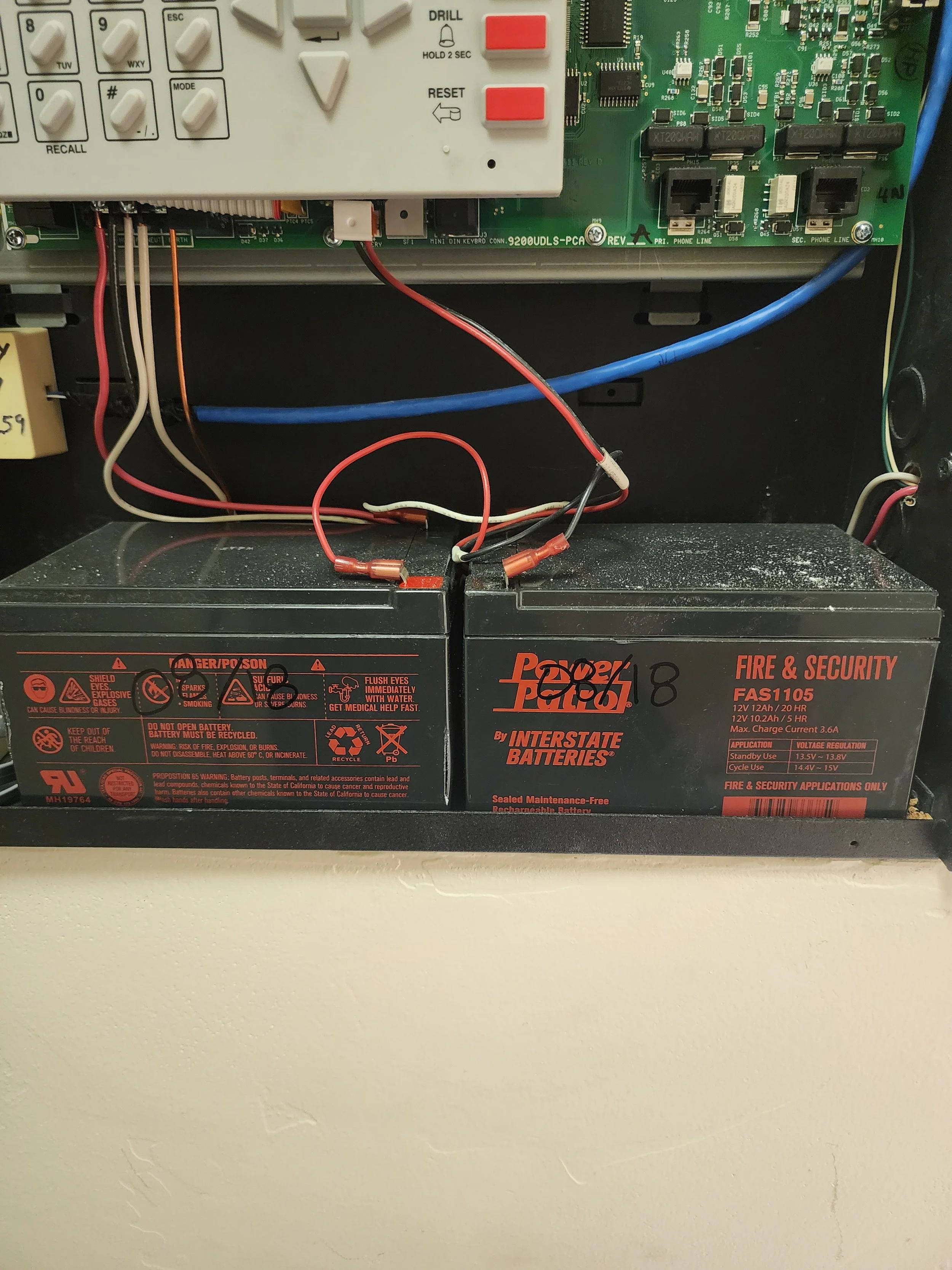 View of a black interstate batteries fire and security backup power supply with wires connected inside a metal enclosure. There is also a circuit board and a white control panel with buttons above.