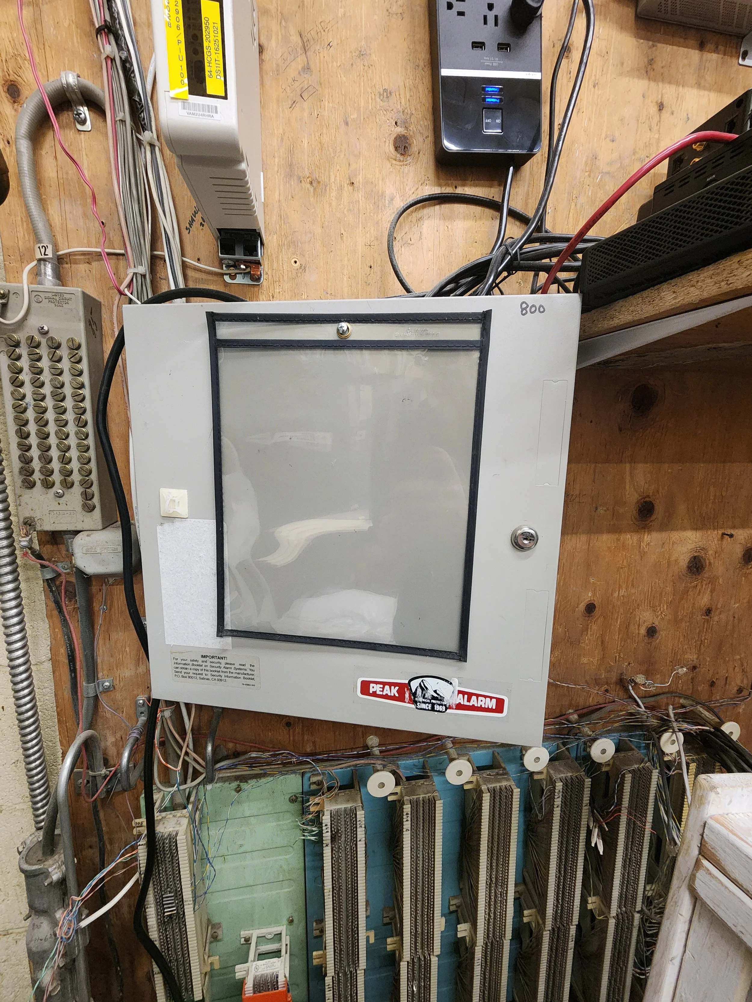 An electrical control panel attached to a wooden wall, with multiple wires, circuit boards, and a PEAK alarm sticker.