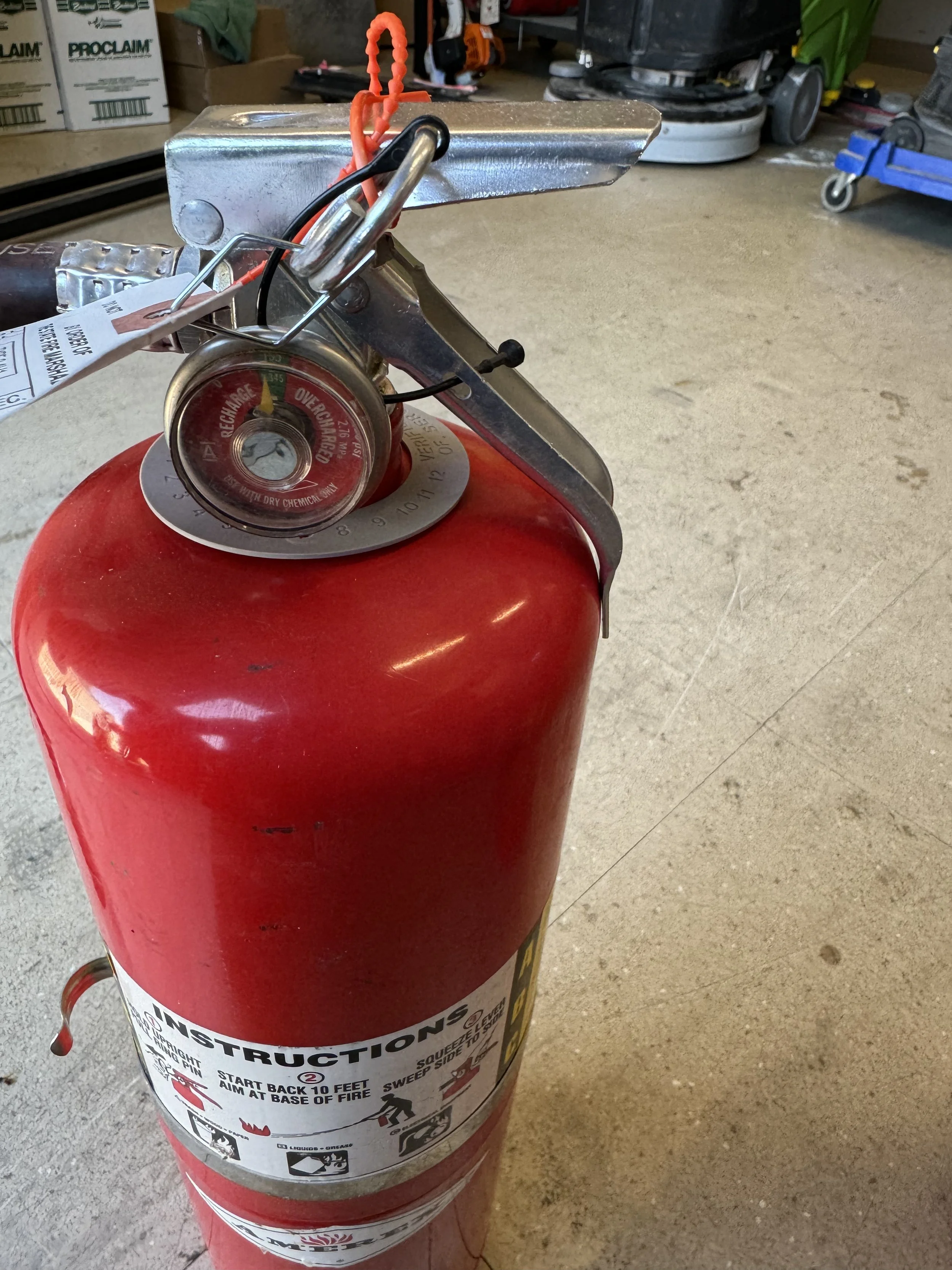 A red fire extinguisher with a pressure gauge and instructions attached, placed on a concrete floor in a workshop or garage.