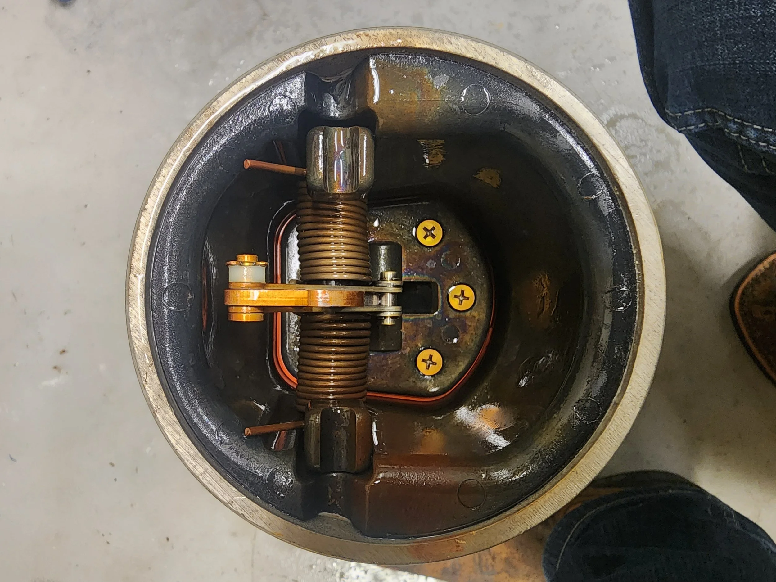 Top view of a mechanical device with a large spring, yellow screws, and metallic components inside a circular housing.