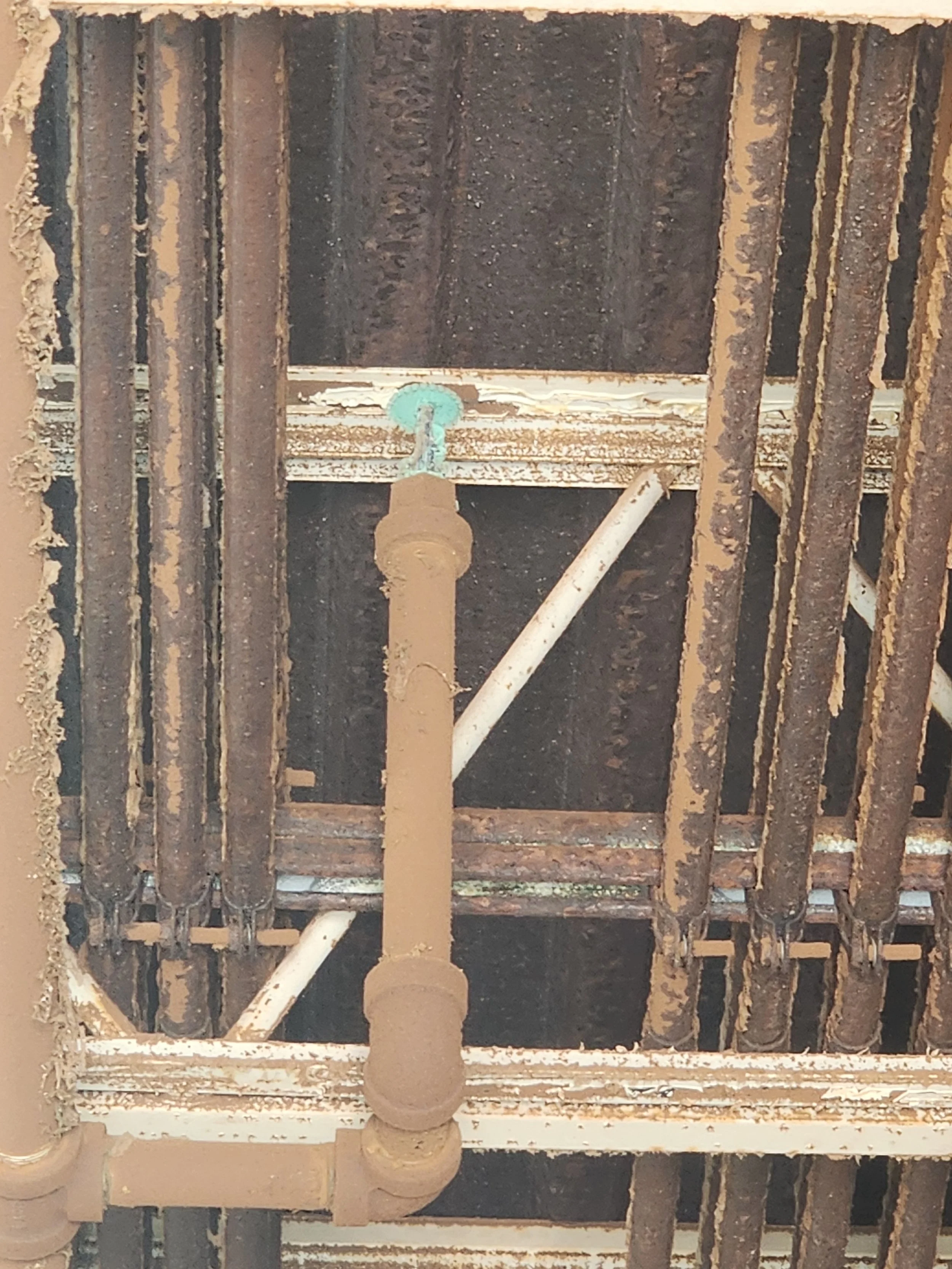A ceiling with old, rusted steel beams, some white pipes, and a brown painted metal pipe with a valve, seen from below.