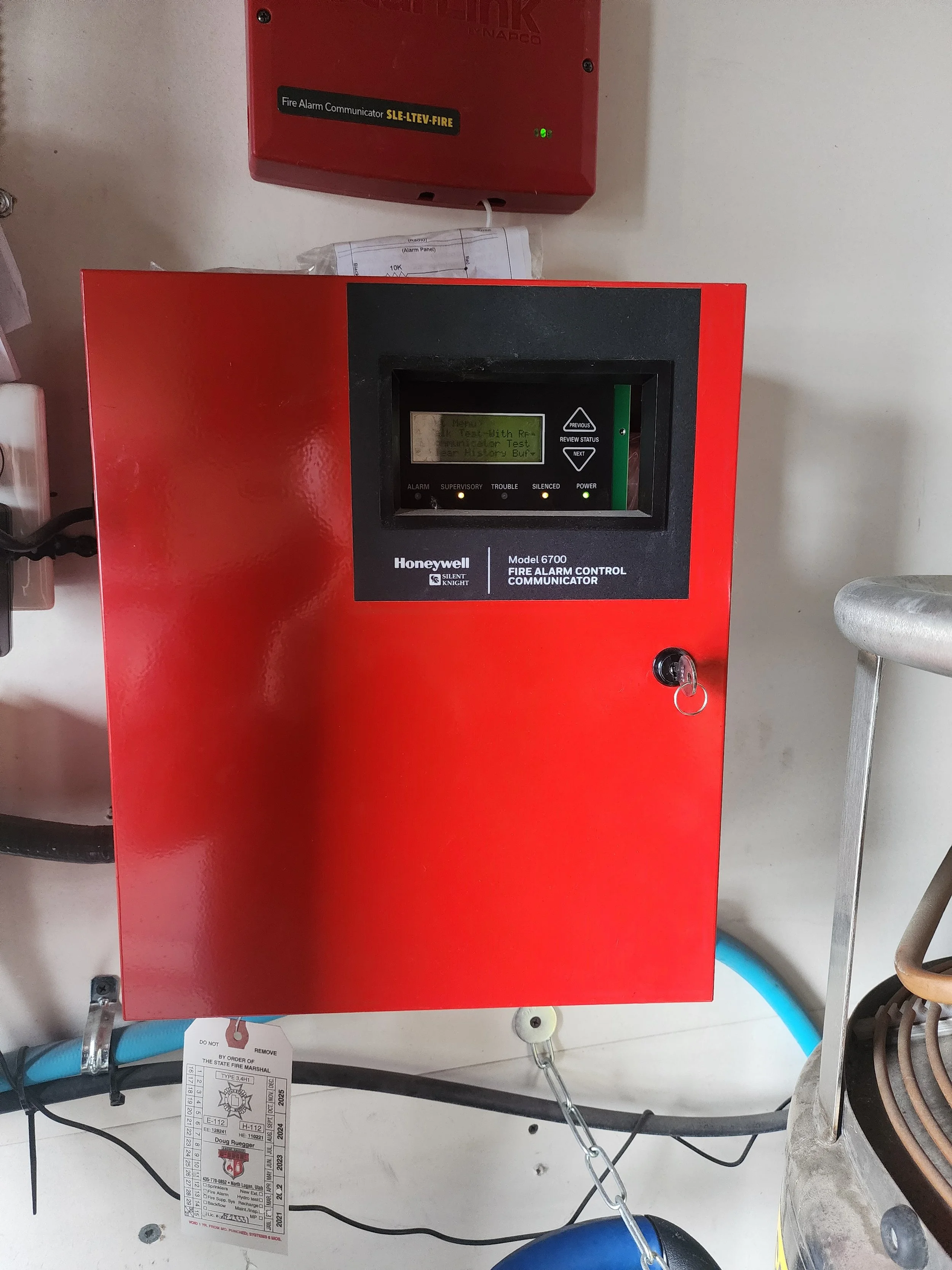 Red Honeywell fire alarm control panel with a small digital display, located on a white wall, with labels for alarm, supervisor, trouble, silenced, and power.