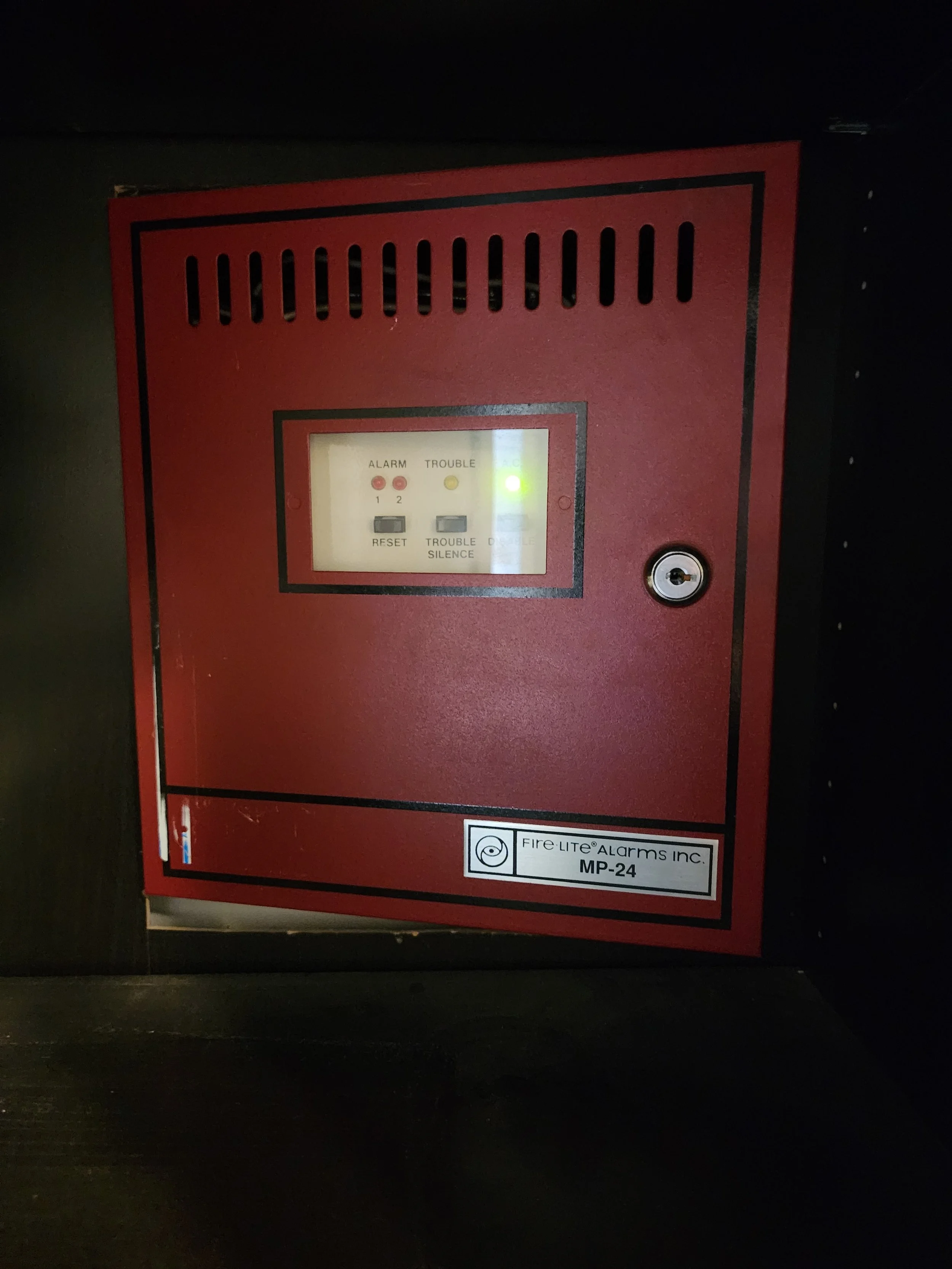 A red fire alarm control panel with indicator lights and buttons installed inside a dark wall cabinet.