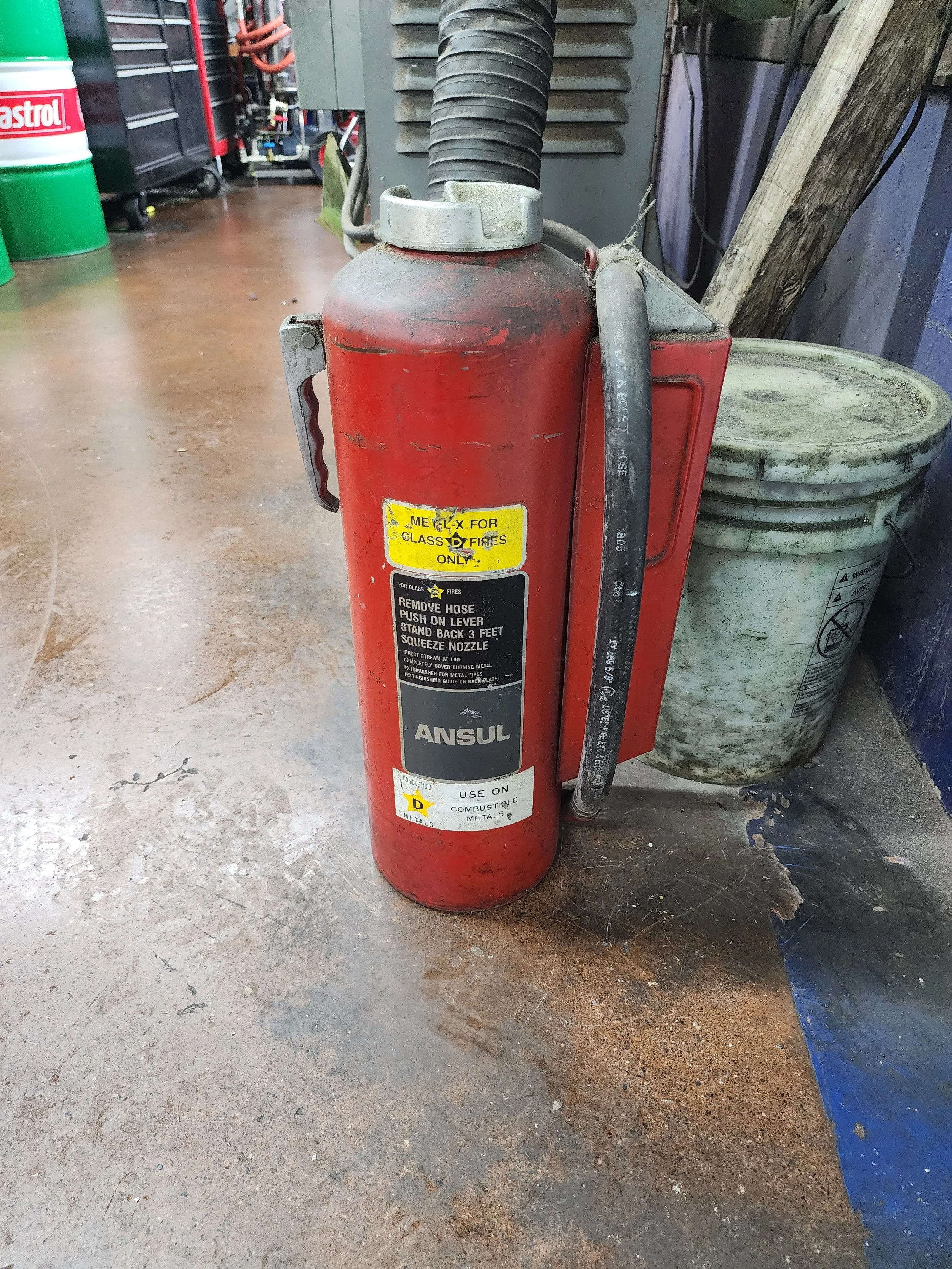 A red fire extinguisher labeled for Class D fires, used on combustible metals, with a hose attached, standing on a concrete floor in a workshop area.