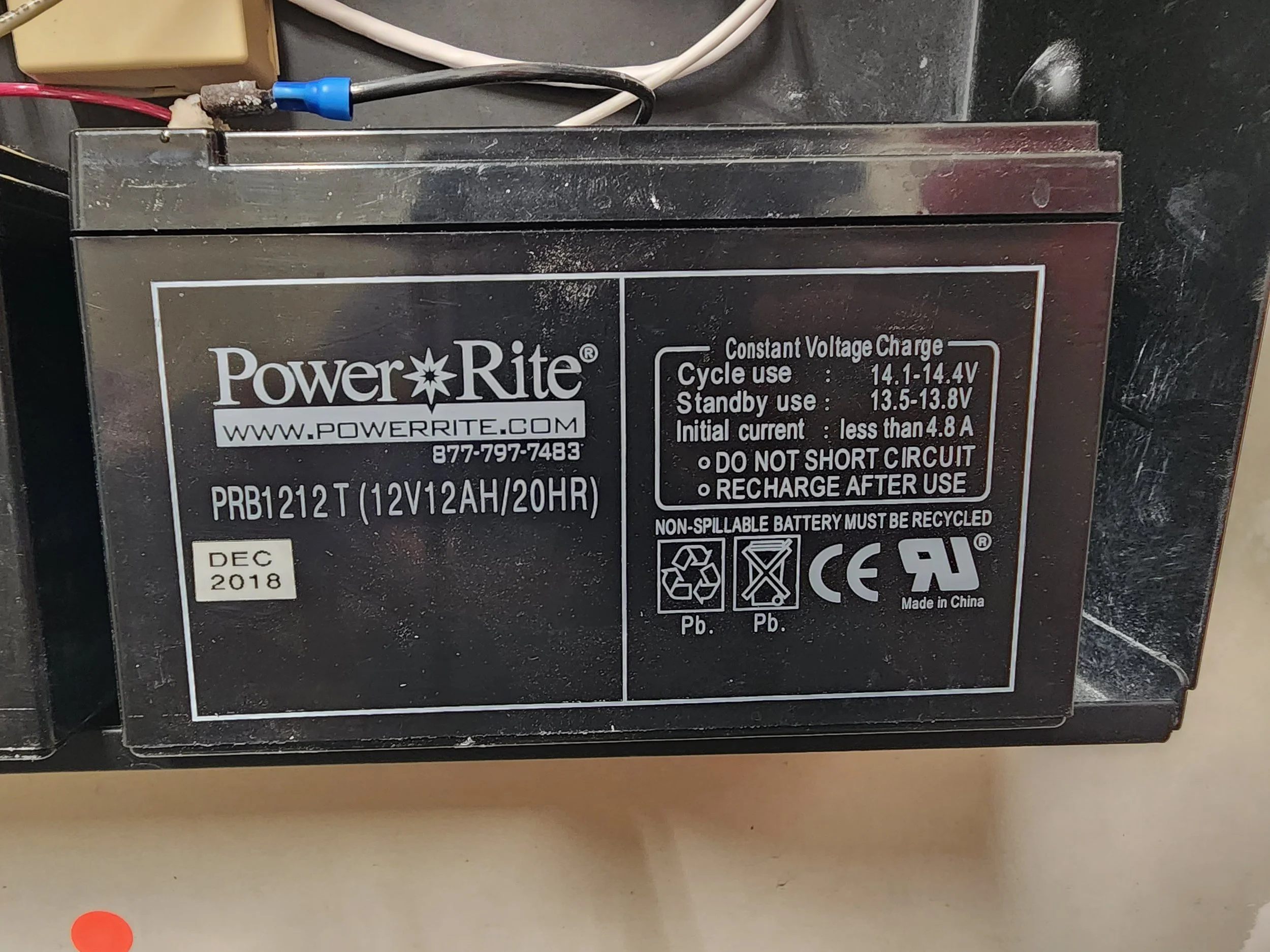 A Power-Rite 12-volt, 12 amp-hour, 20-hour lead-acid battery with a black casing, labels indicating model and specifications, and various safety symbols. The label shows it was manufactured in December 2018 and includes instructions for use and recyc