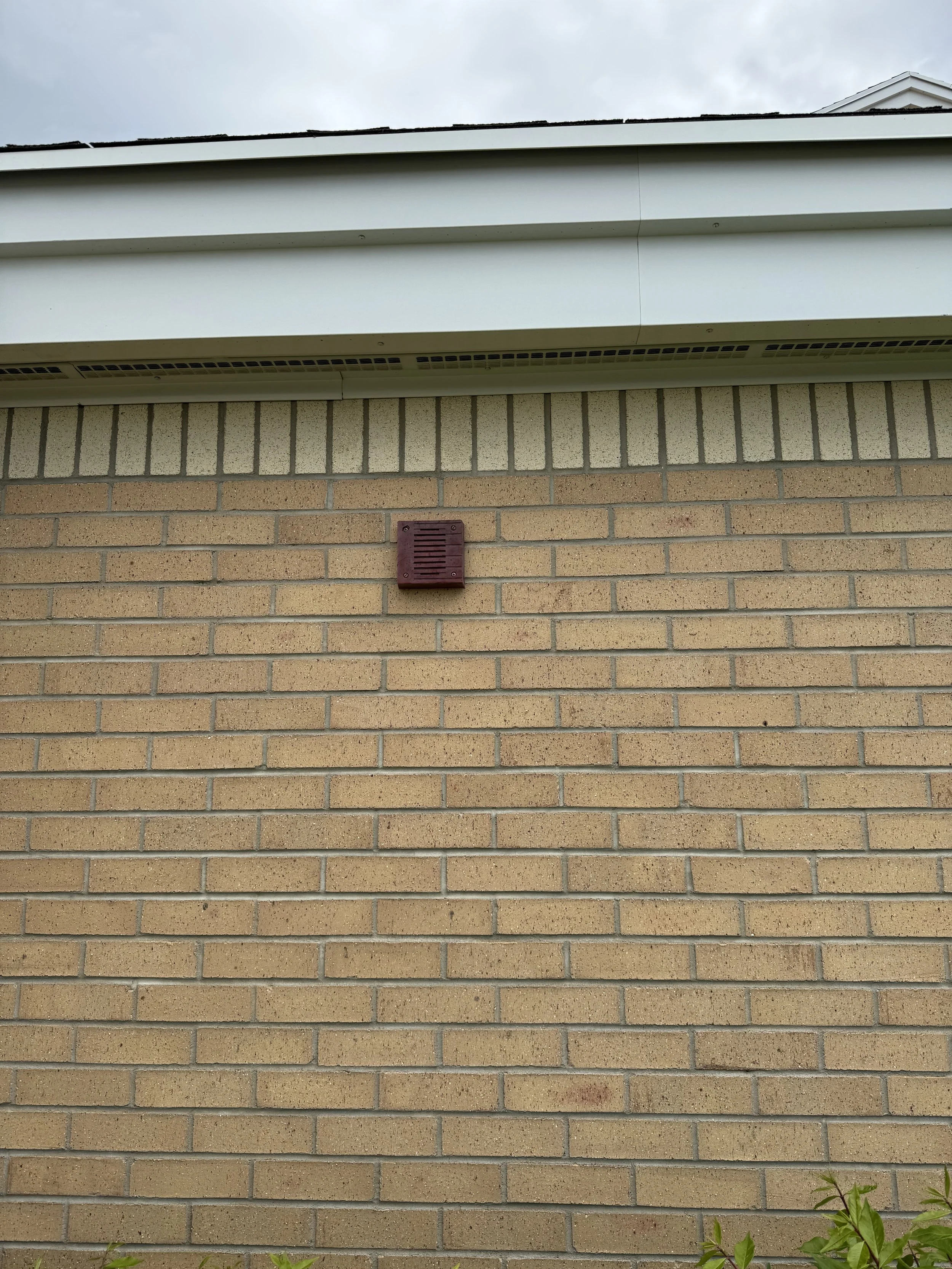 Close-up of a beige brick building wall with a small red vent near the center and white siding at the top, with an overcast sky above.