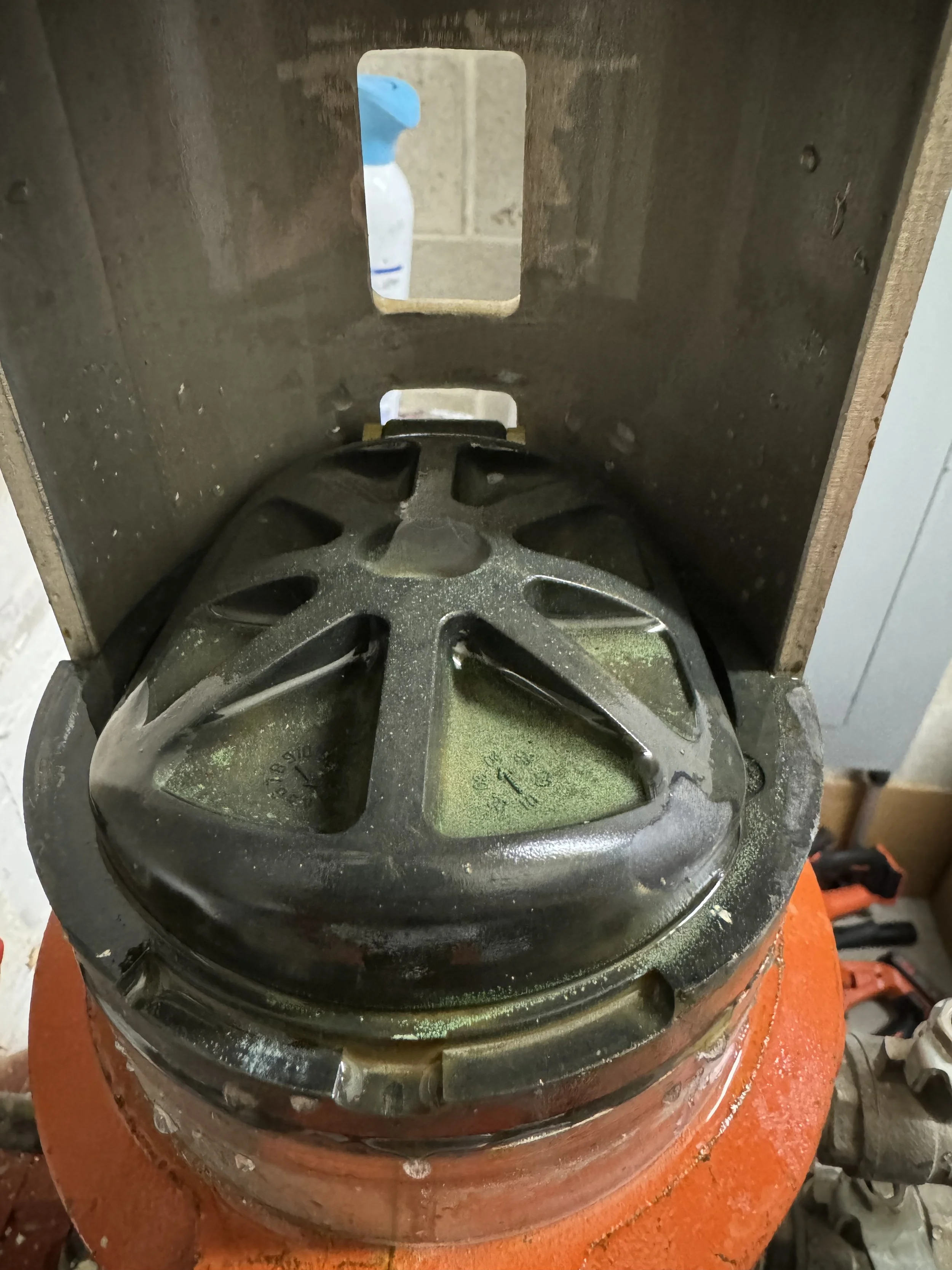 A speaker mounted inside a wooden box, with a metal grille covering the cone, placed on an orange surface in a workshop setting.