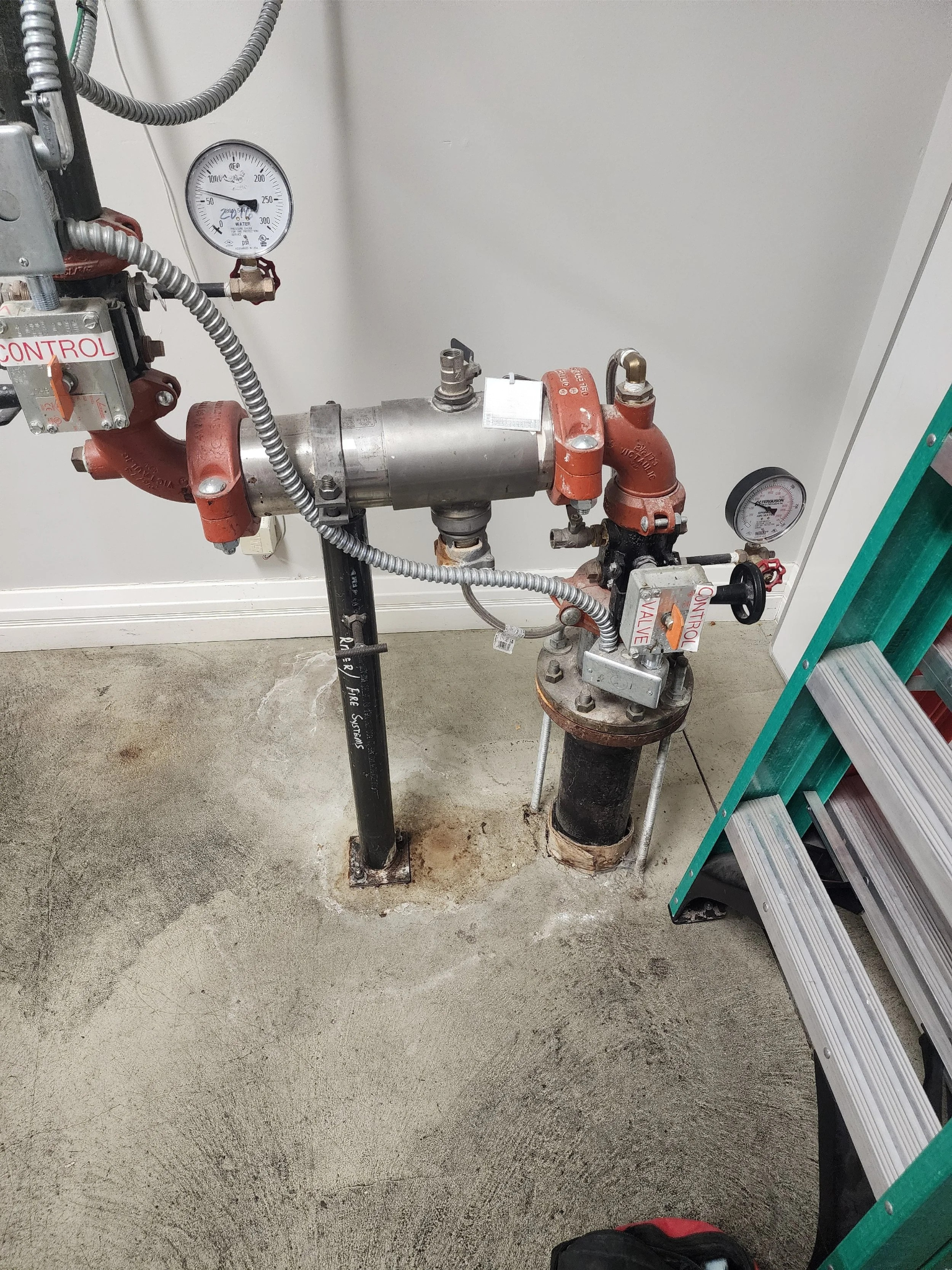 A fire sprinkler system pipe with control valves and pressure gauges, installed near a corner of a concrete floor and wall, with a ladder nearby.