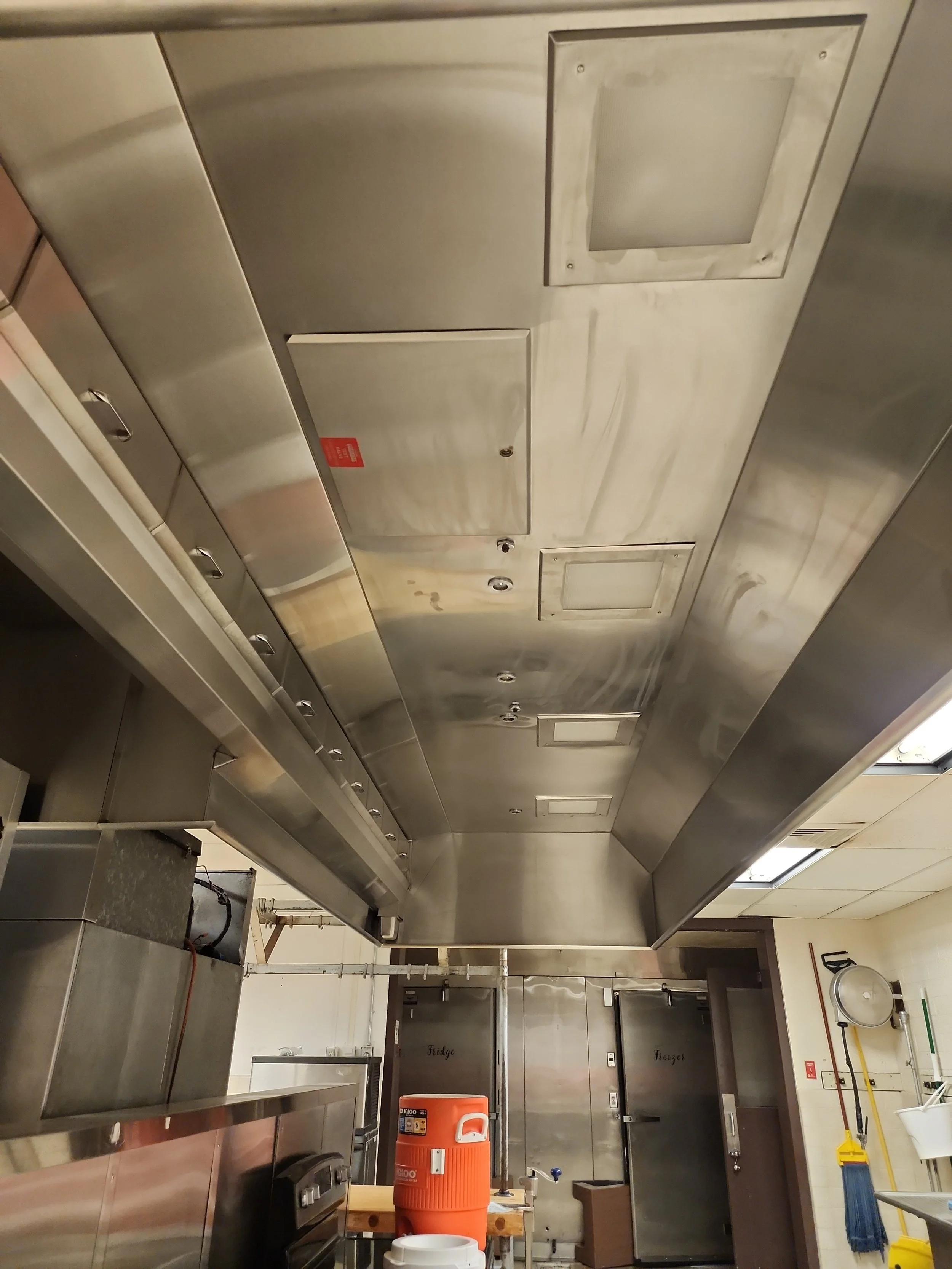Industrial kitchen with stainless steel surfaces, large commercial refrigerators, and cleaning supplies visible on the side.