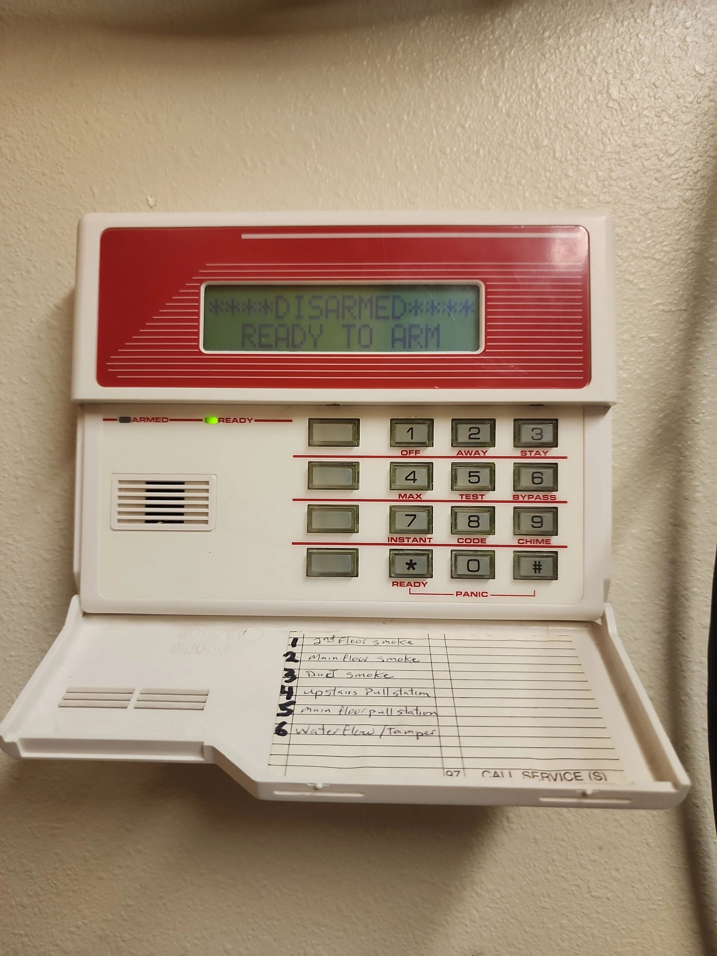 Fire alarm control panel with a note attached below it. The display reads 'DISARMED READY TO ARM'. The note lists several items, including smoke in different floors, upstairs pull station, main floor pull station, and water flow tamper.