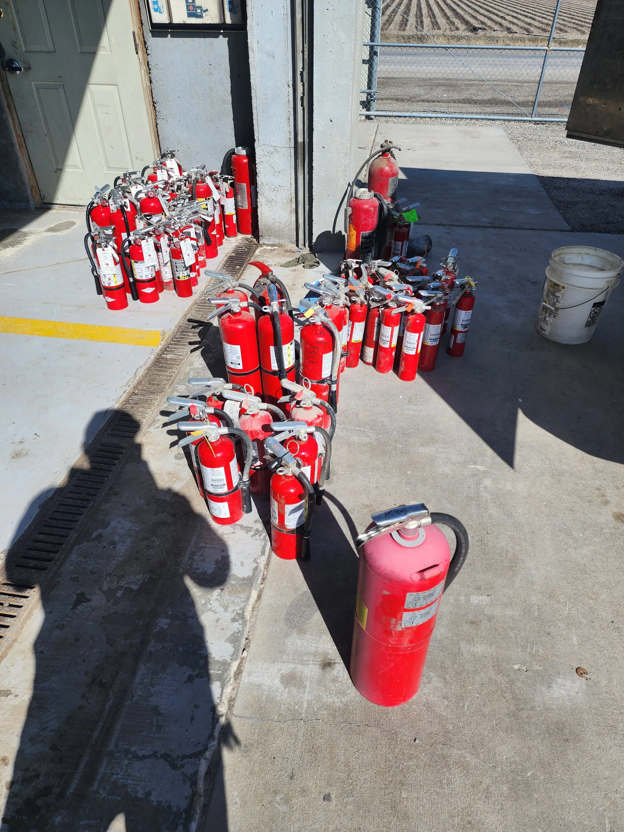 Various fire extinguishers arranged outside a building near a garage door, with some placed on the ground and others on a small shelf.