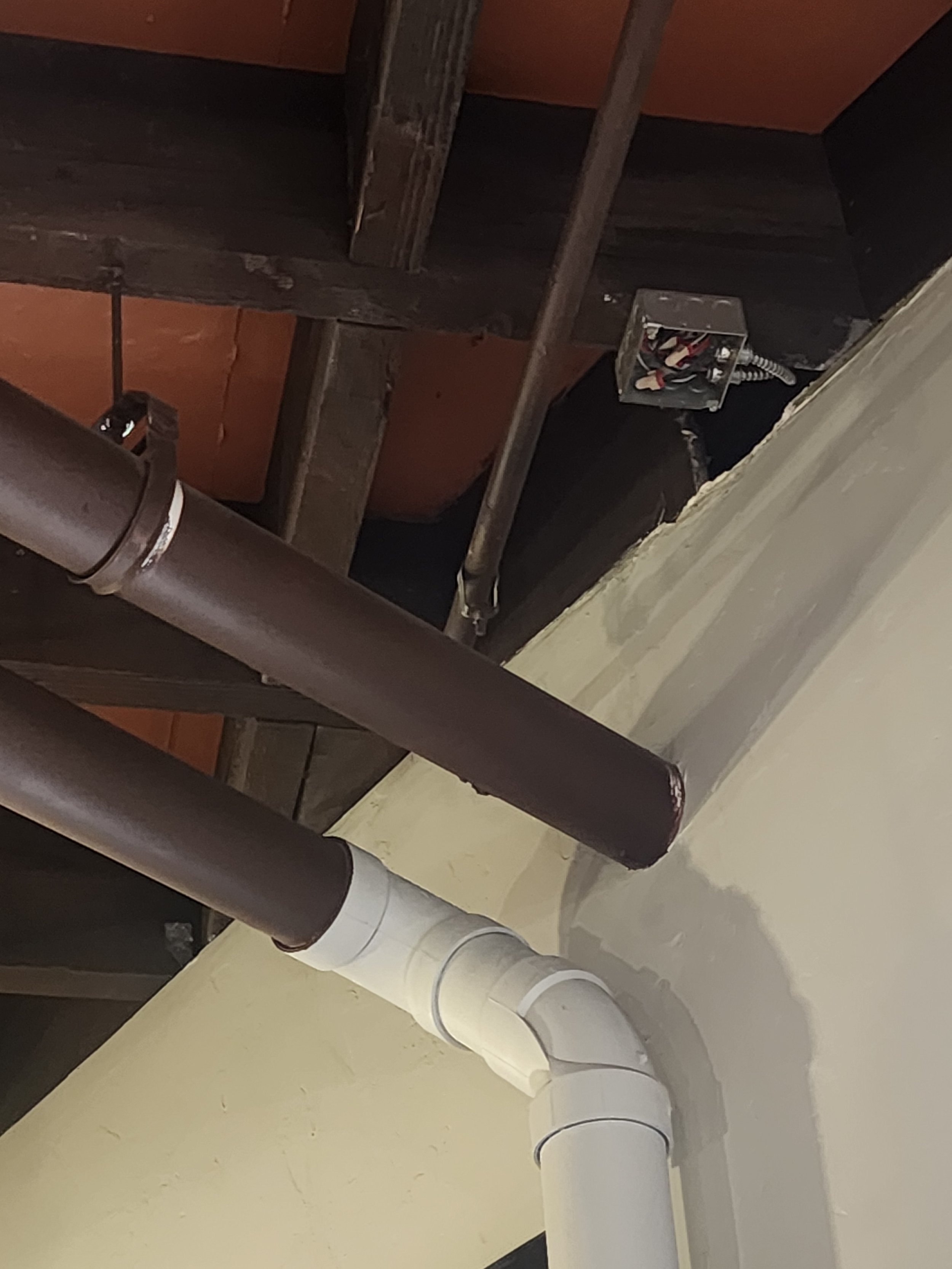 View of plumbing pipes, including a brown pipe and white pipe, running along a corner of a room under construction with exposed ceiling beams and an electrical box.