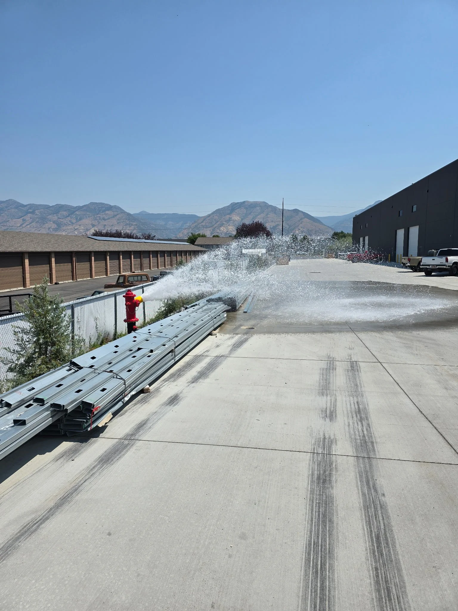 A fire hydrant releasing a powerful stream of water onto a concrete surface outside a warehouse, with a clear blue sky and mountains in the background.