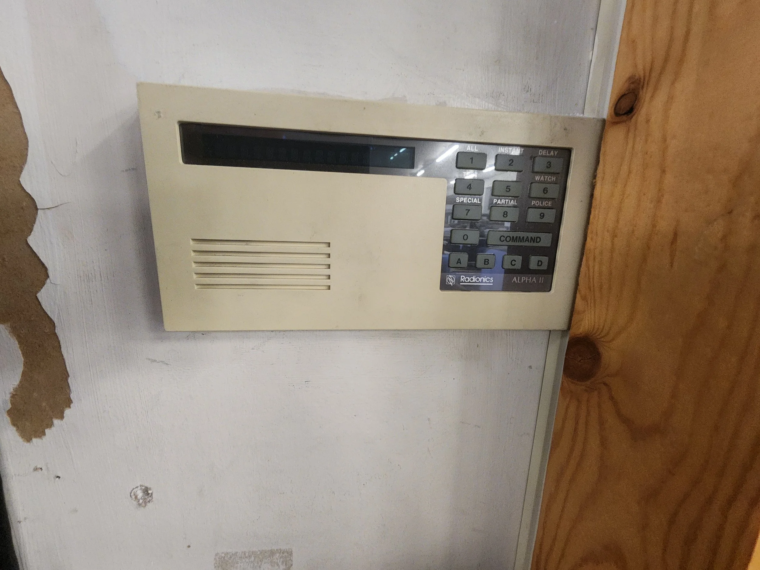 An old beige intercom or security alarm system mounted on a wall with a vent and buttons for various functions.