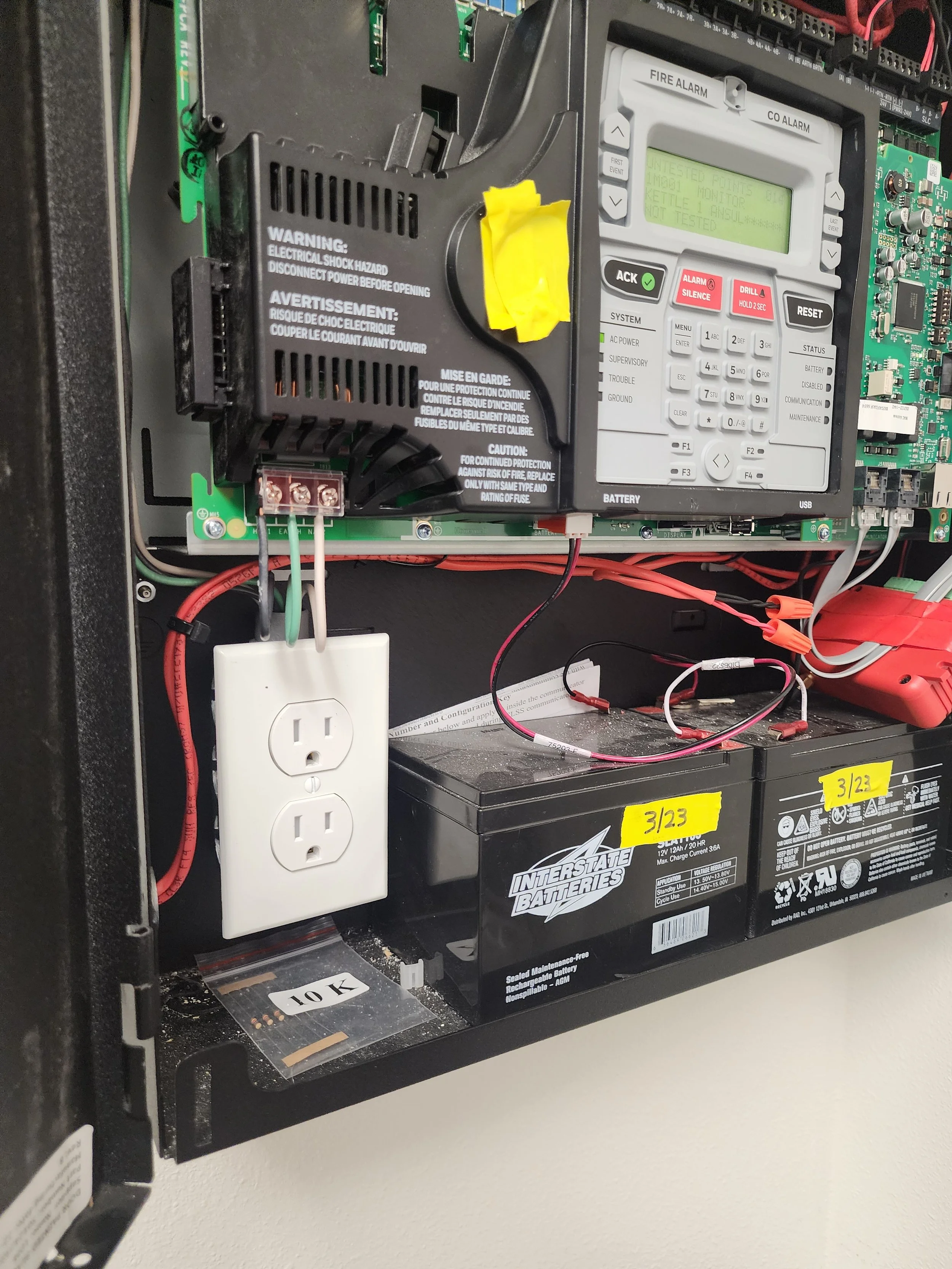 Electrical control panel with a black circuit breaker, a digital timer switch, and two batteries labeled with yellow stickers '3/23' and '3/24'.