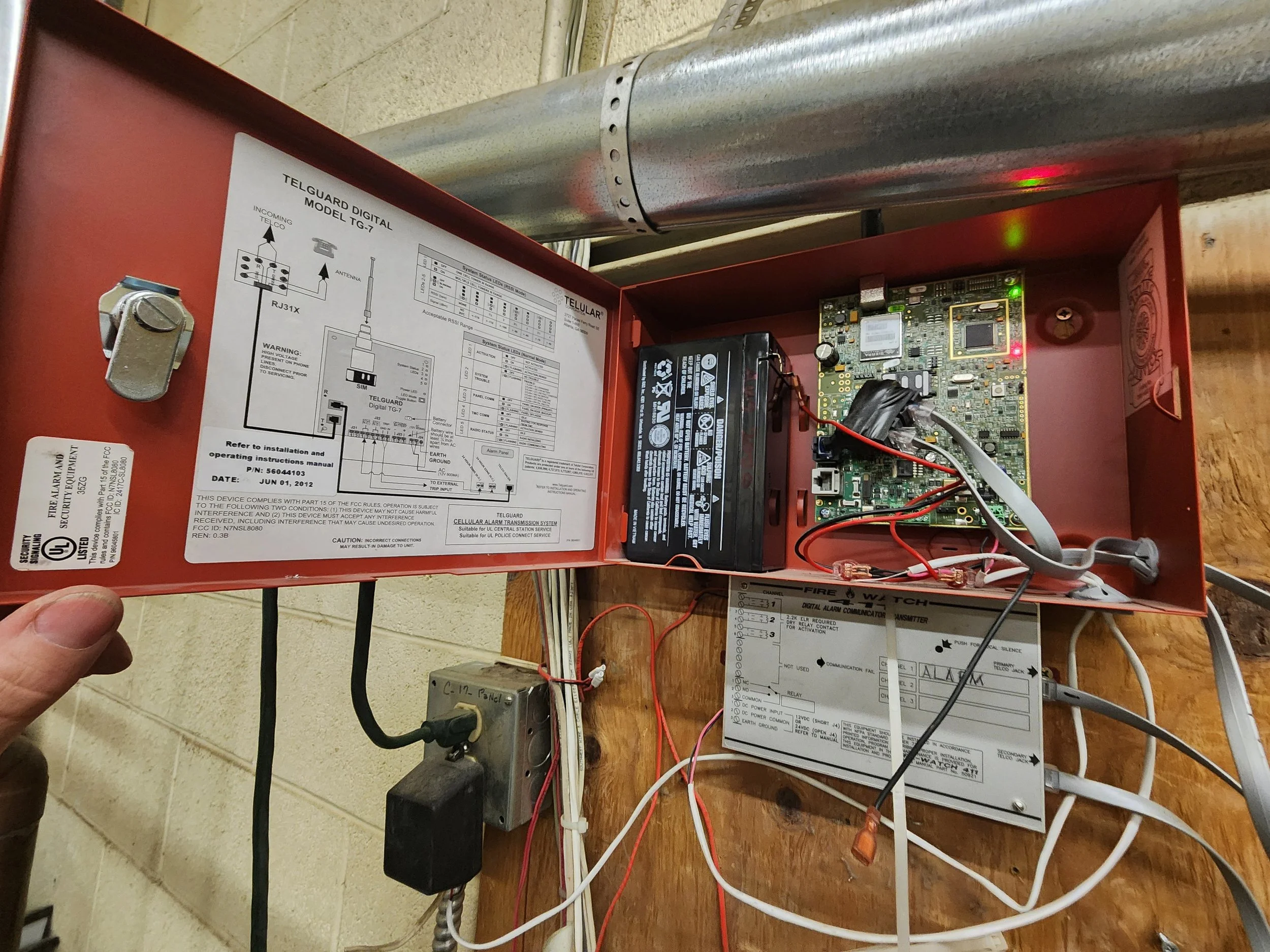 Open red metal box containing telecommunication and security system components, including circuit boards, a black battery, wiring, and a diagram on the inside of the lid.
