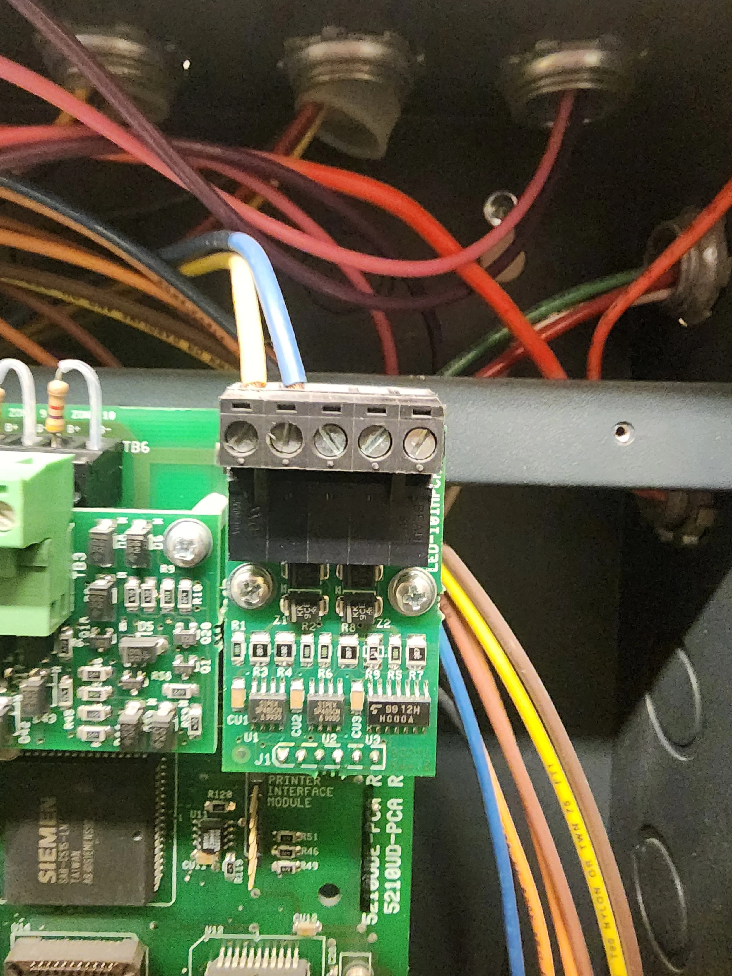 Close-up of a green printed circuit board with various electronic components and wires connected to a terminal block, inside an electronic device or machine.