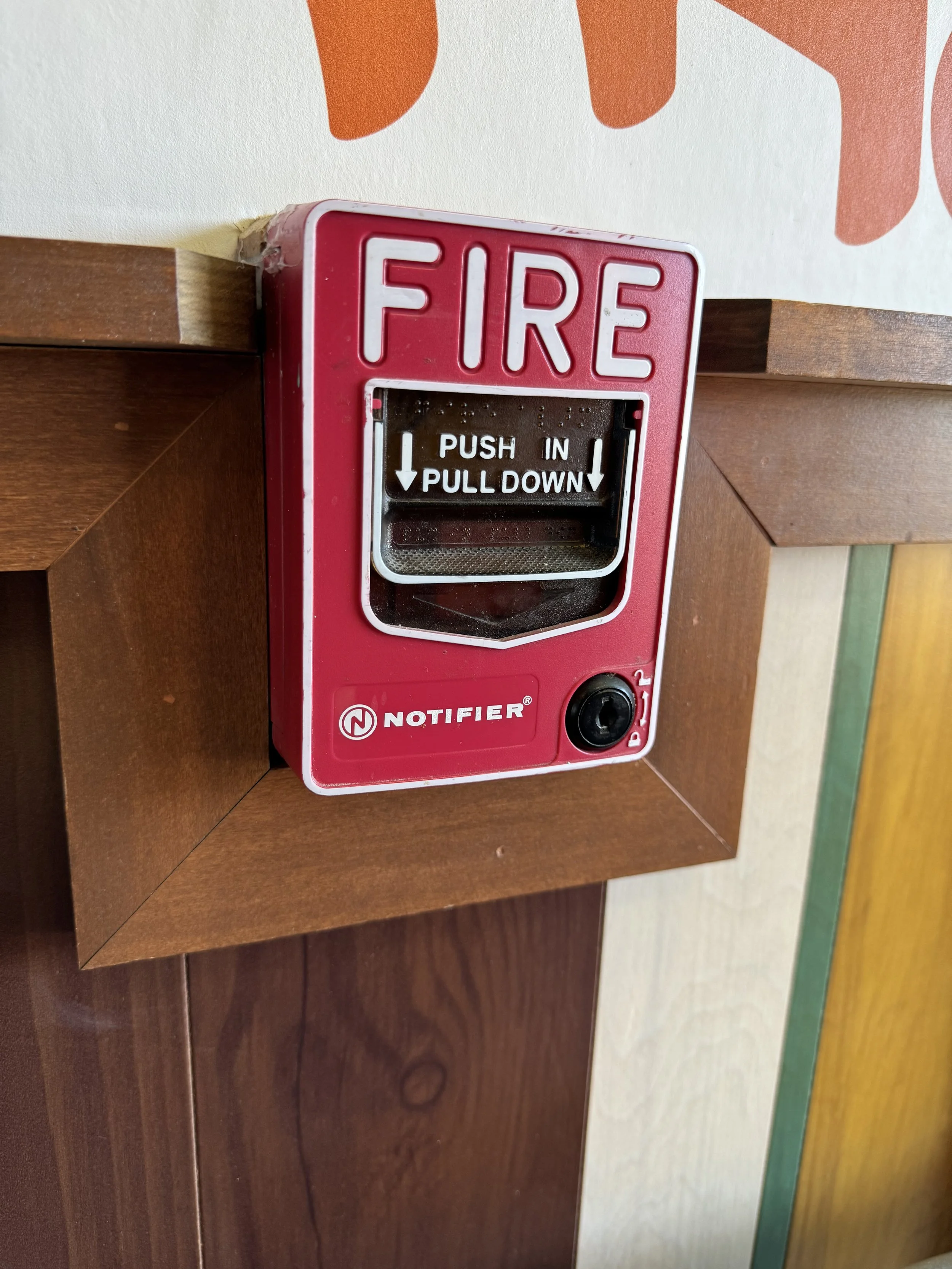 A red fire alarm pull station mounted on a wooden wall with gold and green trim. The station has a label that says 'FIRE' and instructions to push in and pull down.