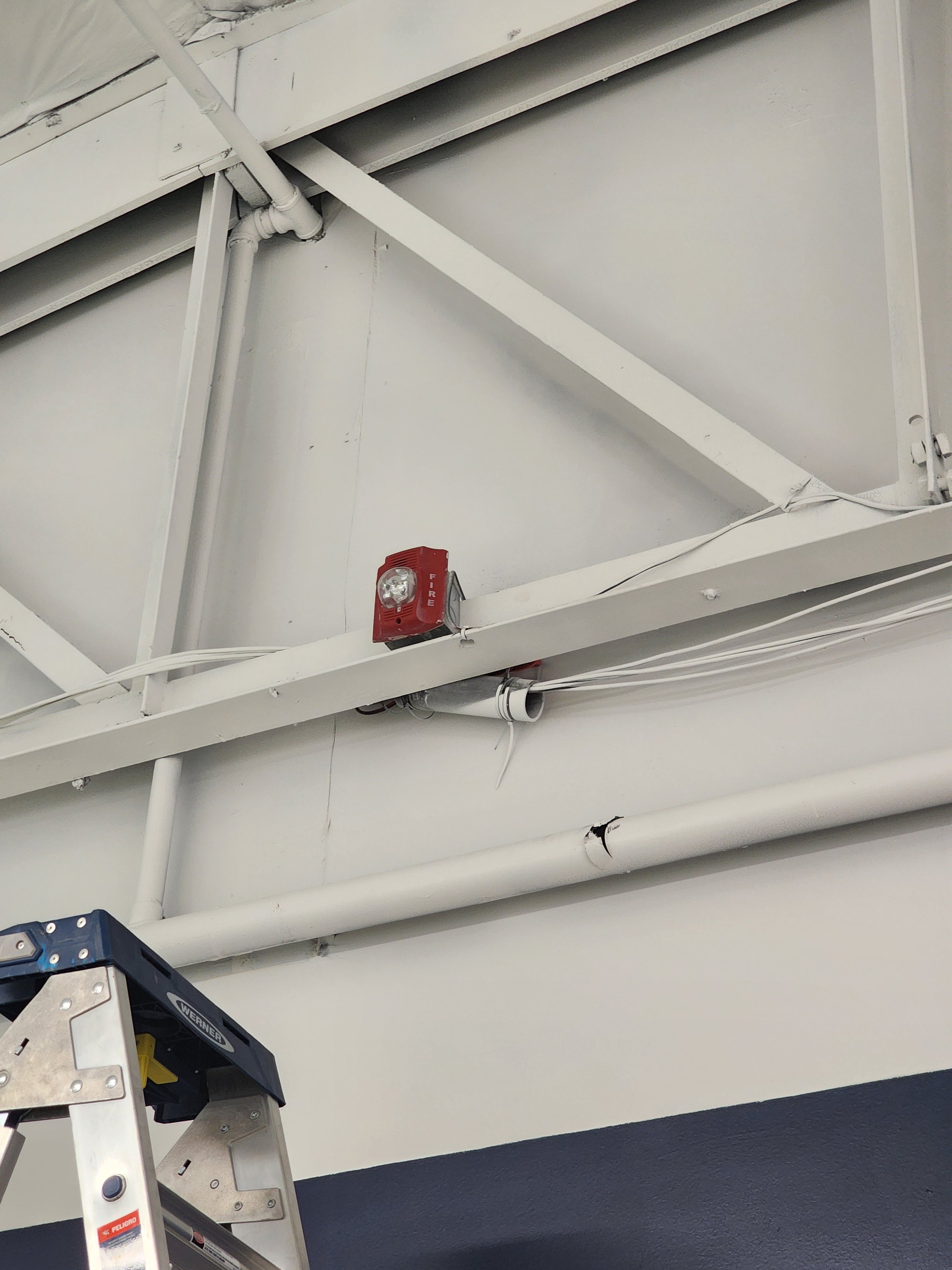 Ceiling with exposed pipes, a red alarm, and a partially visible step ladder.