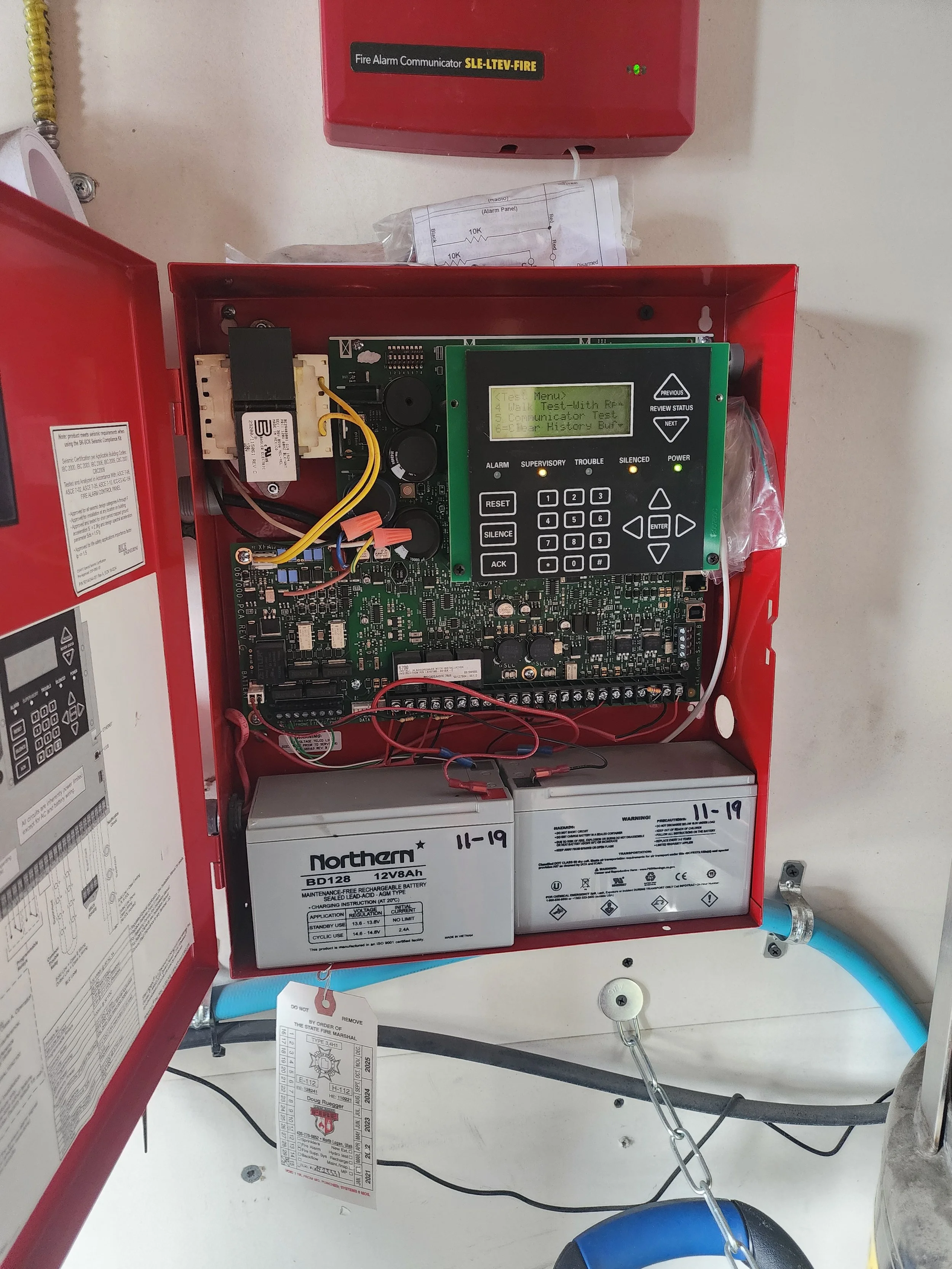 Open fire alarm control panel with circuit board, batteries, and wiring inside, mounted on a wall.
