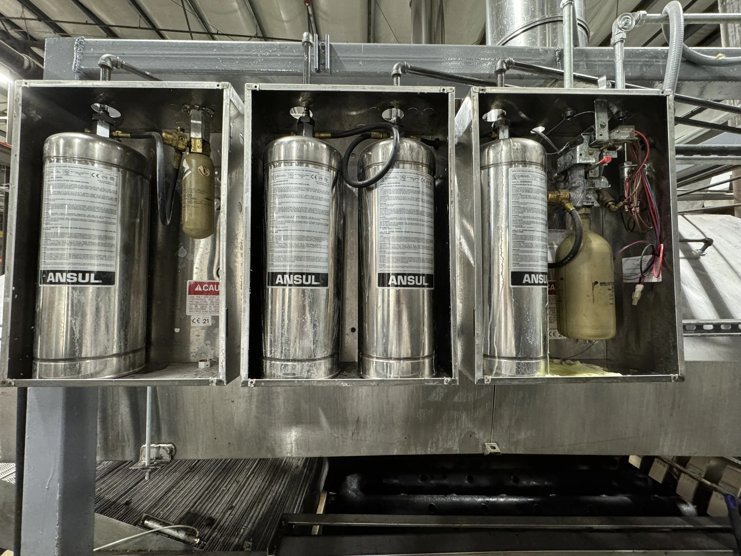 Four stainless steel cylinders labeled ANSUL inside a metal industrial cabinet, with pipes and wires connected.