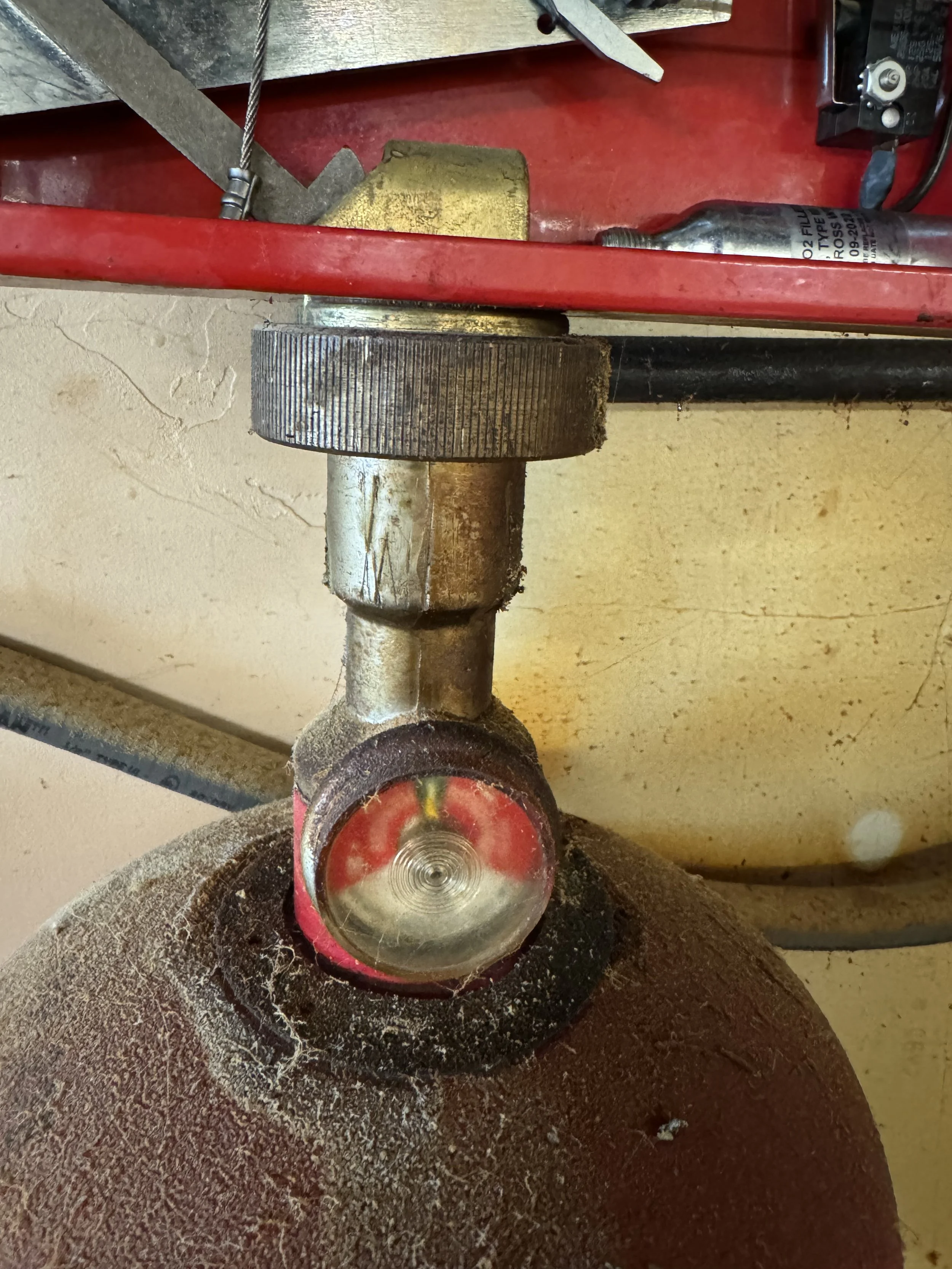 Close-up view of a fire extinguisher attached to a red metal shelf, with the nozzle pointing downward. The nozzle is metallic with a red protective cap, and there is dusty dirt on the surface of the extinguisher.