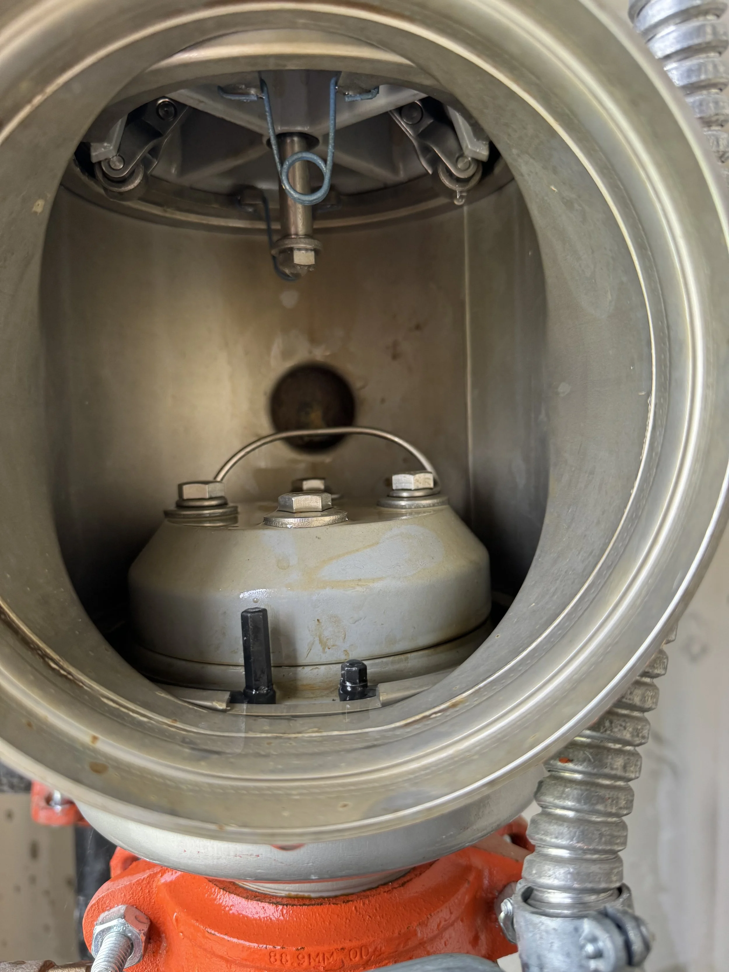 Looking inside an industrial or mechanical component, showing a metallic interior with a circular opening, bolts, a metal arm or pipe at the top, and an orange base at the bottom.