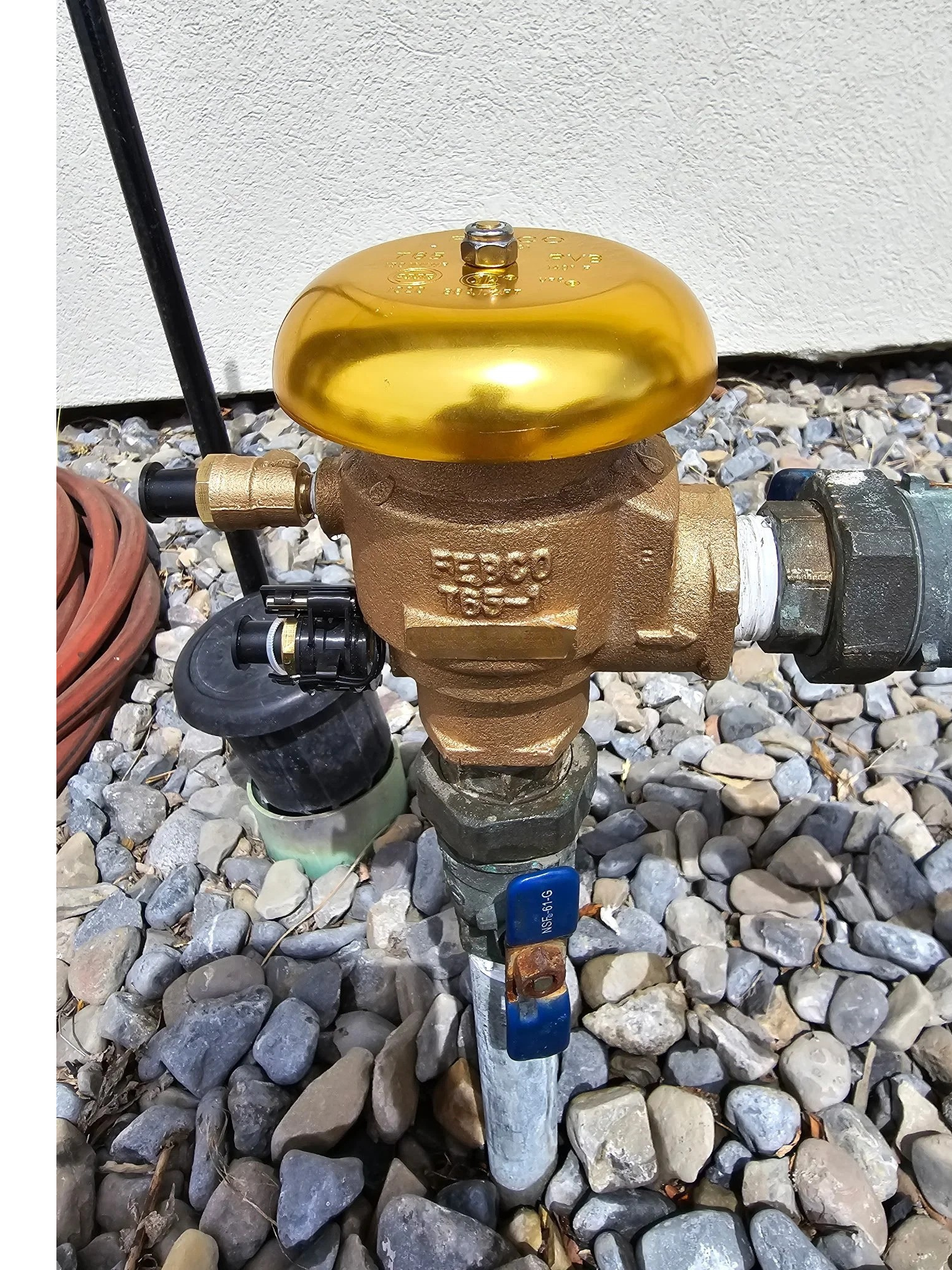 Close-up of a brass sprinkler valve with a blue handle, installed outdoors on a pipe, surrounded by small gray and tan rocks.