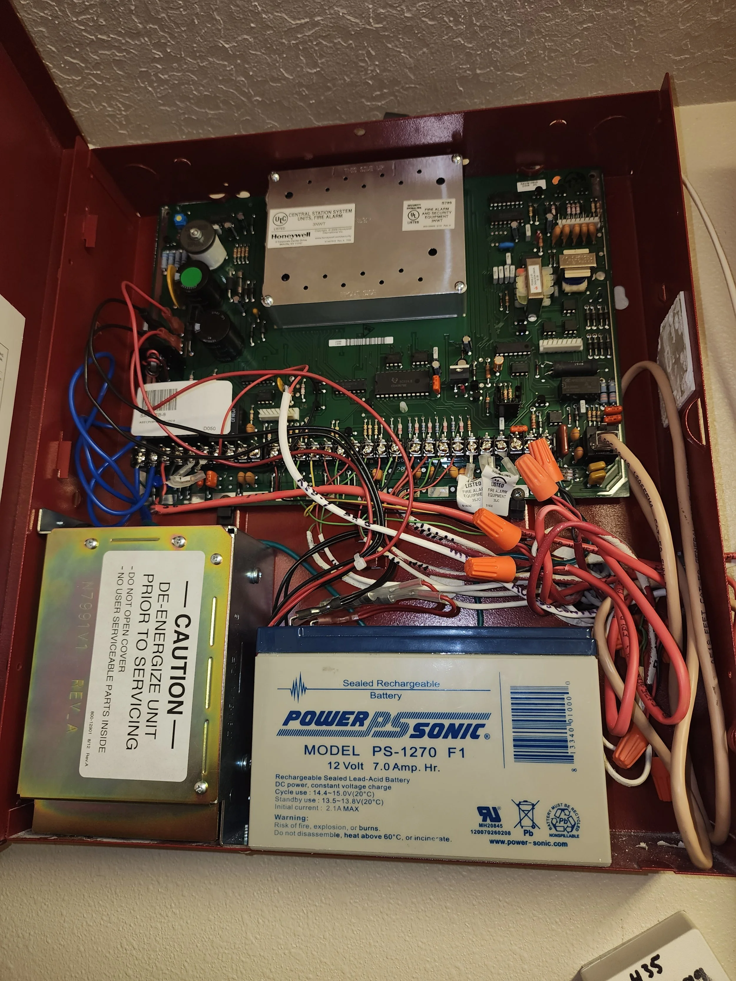 Interior view of a fire alarm control panel with various wiring and electronic components, including a sealed rechargeable battery and power supply module.