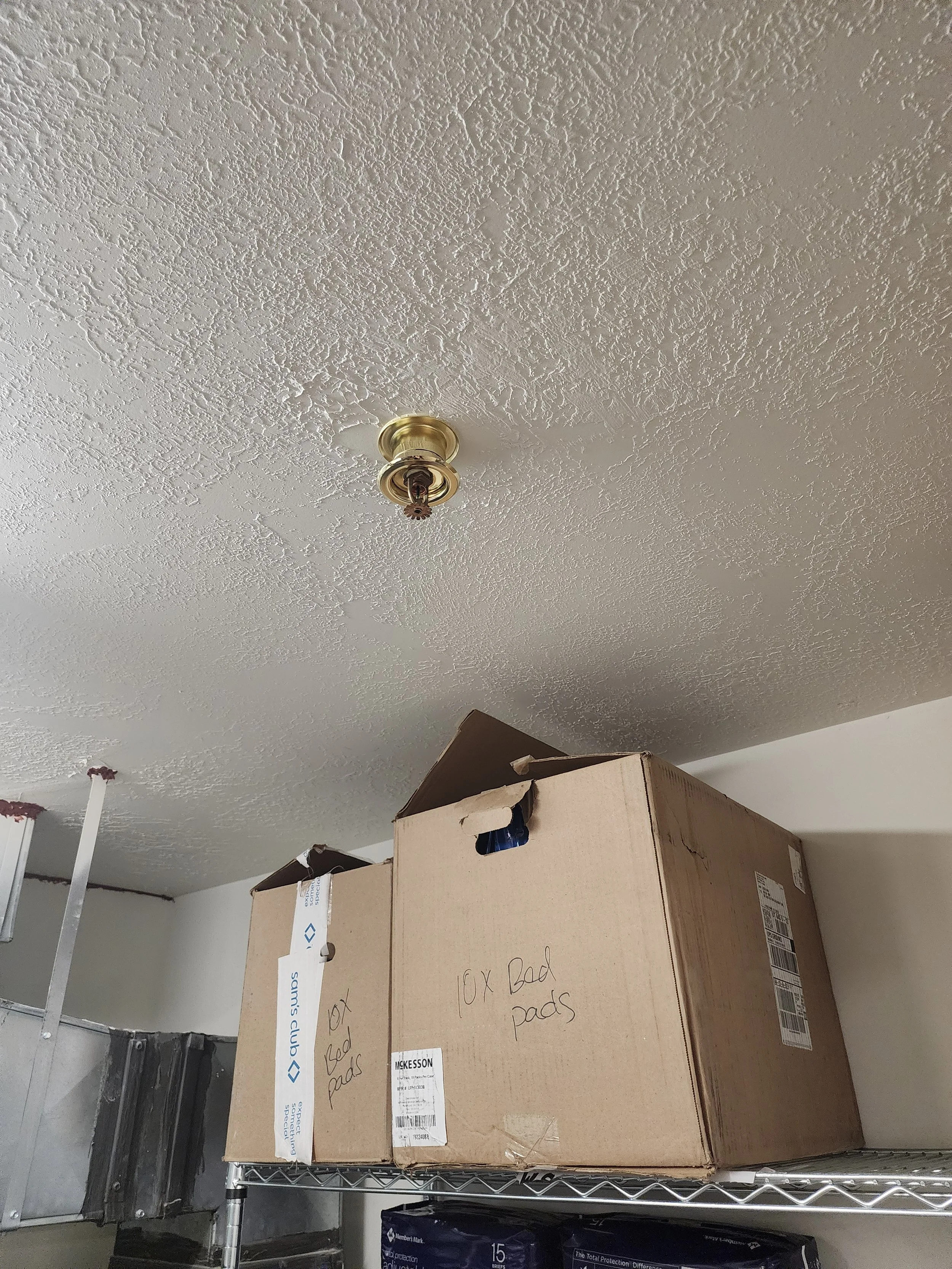 Cardboard boxes labeled "10x Bed Pads" stacked on a metal shelf in a room with a textured ceiling and a missing light fixture.