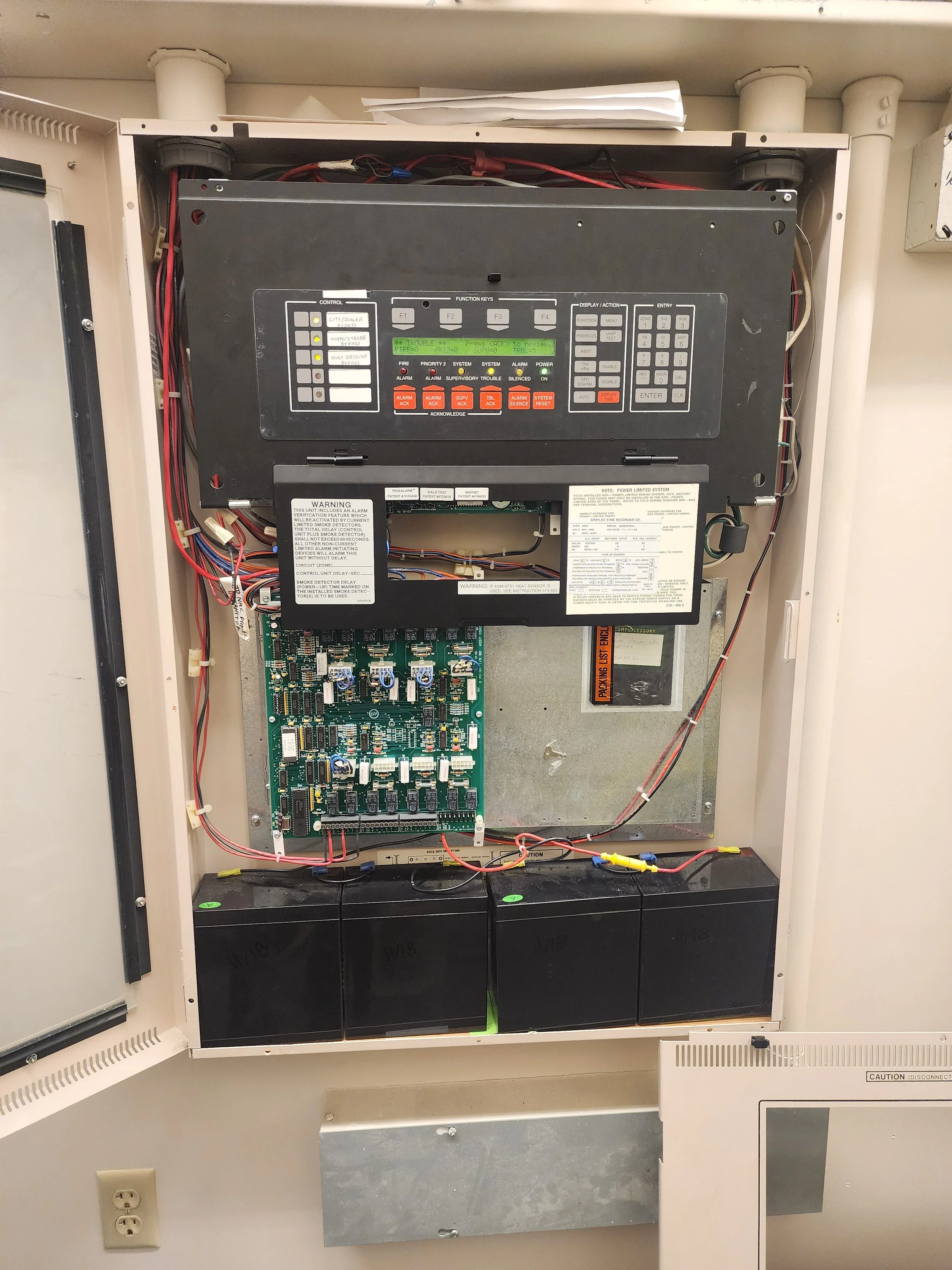 An open fire alarm control panel with various labeled buttons, wires, circuit boards, and batteries inside.