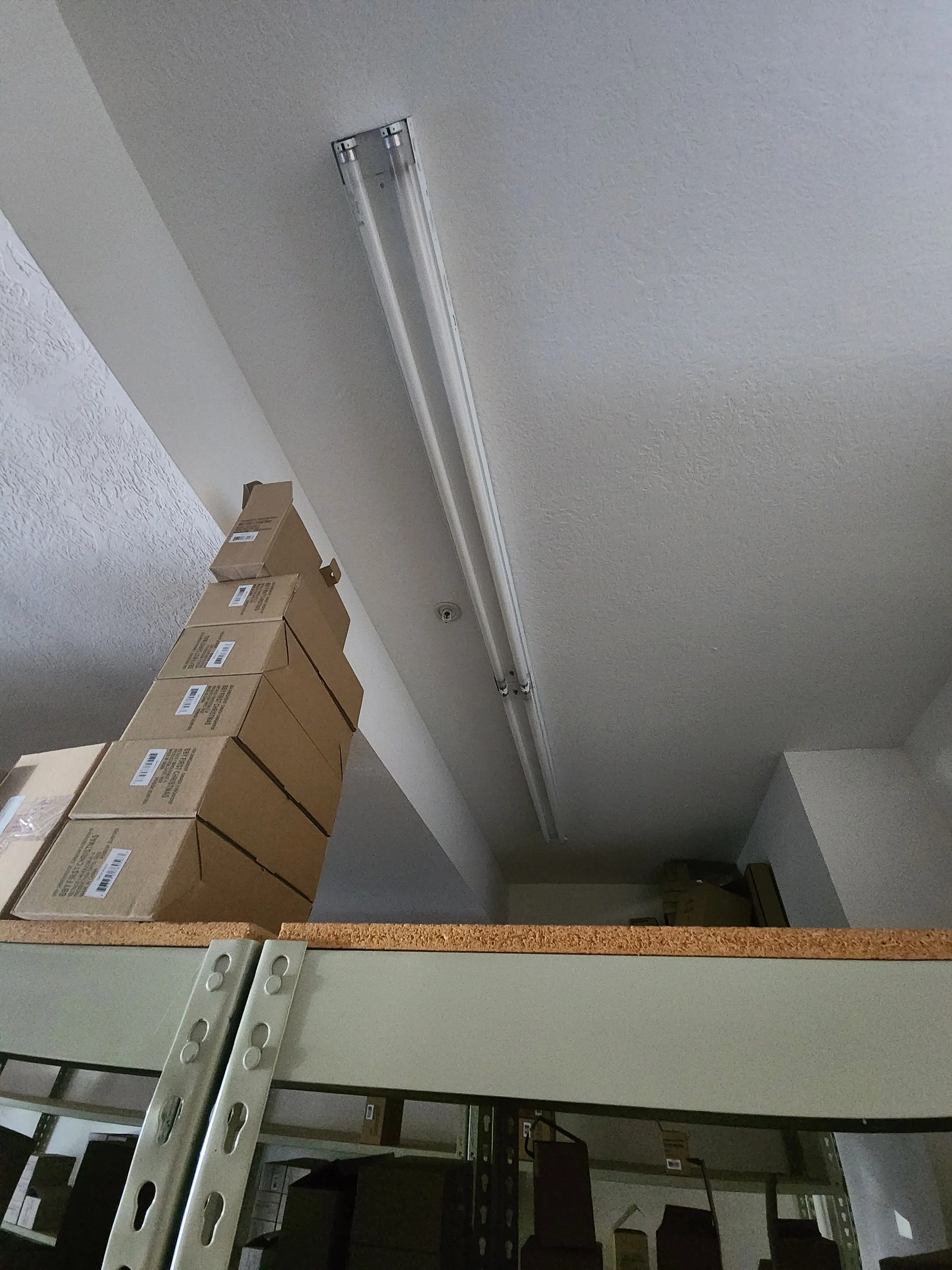 Ceiling with fluorescent light fixture, stacked cardboard boxes on a shelf, and part of a metal shelving unit.