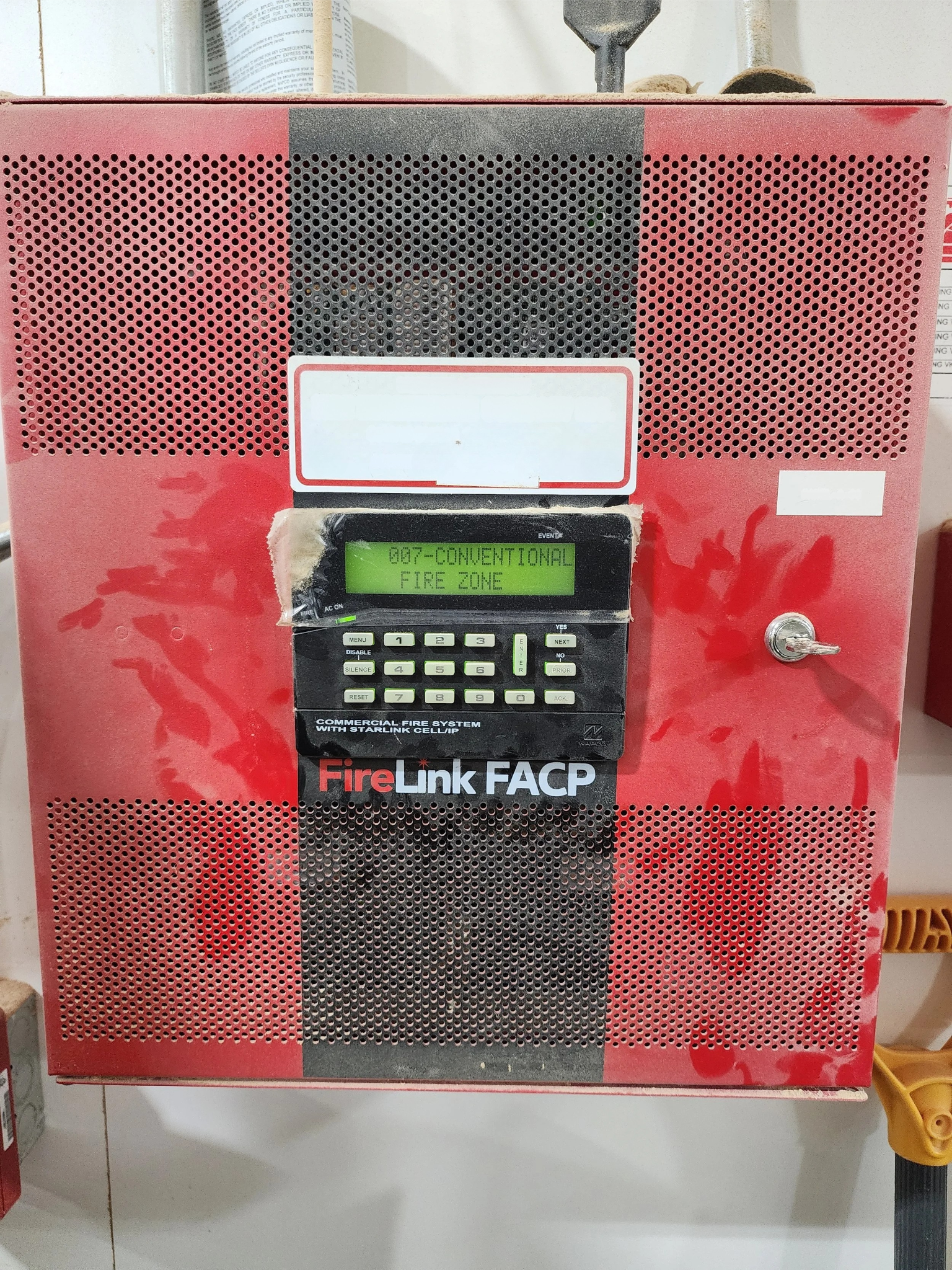 Red fire alarm control panel with a digital display showing '007 CONVENTIONAL FIRE ZONE' and keypad buttons.